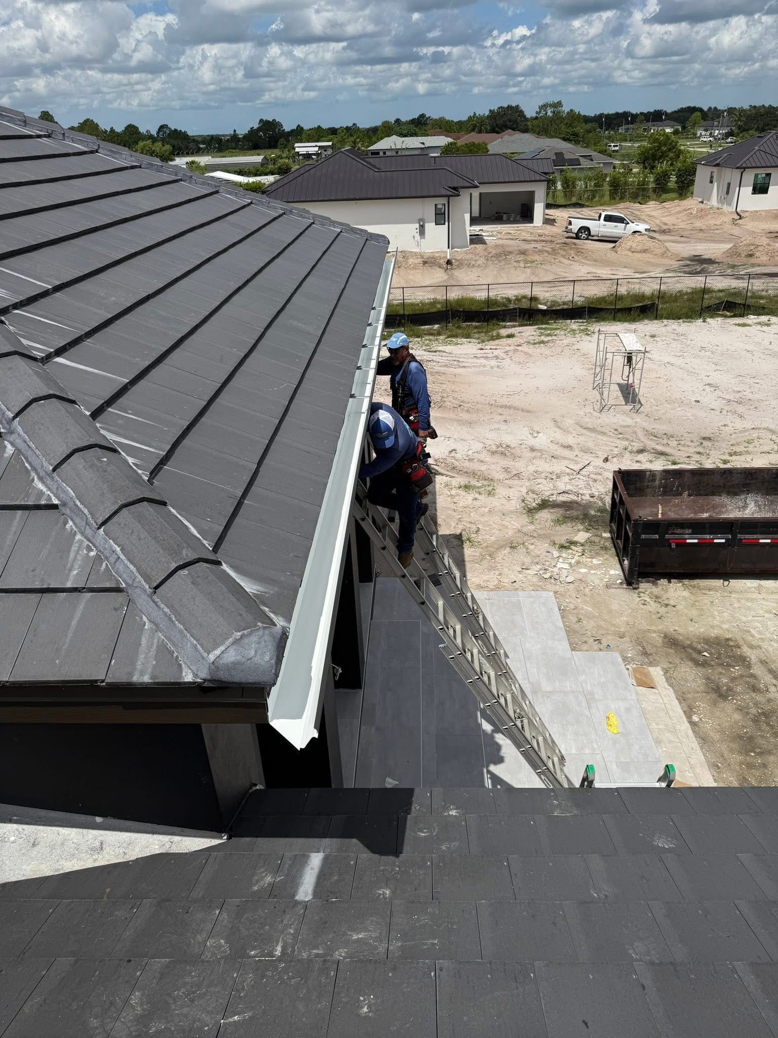 Roofing workers on a gray shingle roof beside a home under construction, with a dirt lot below.