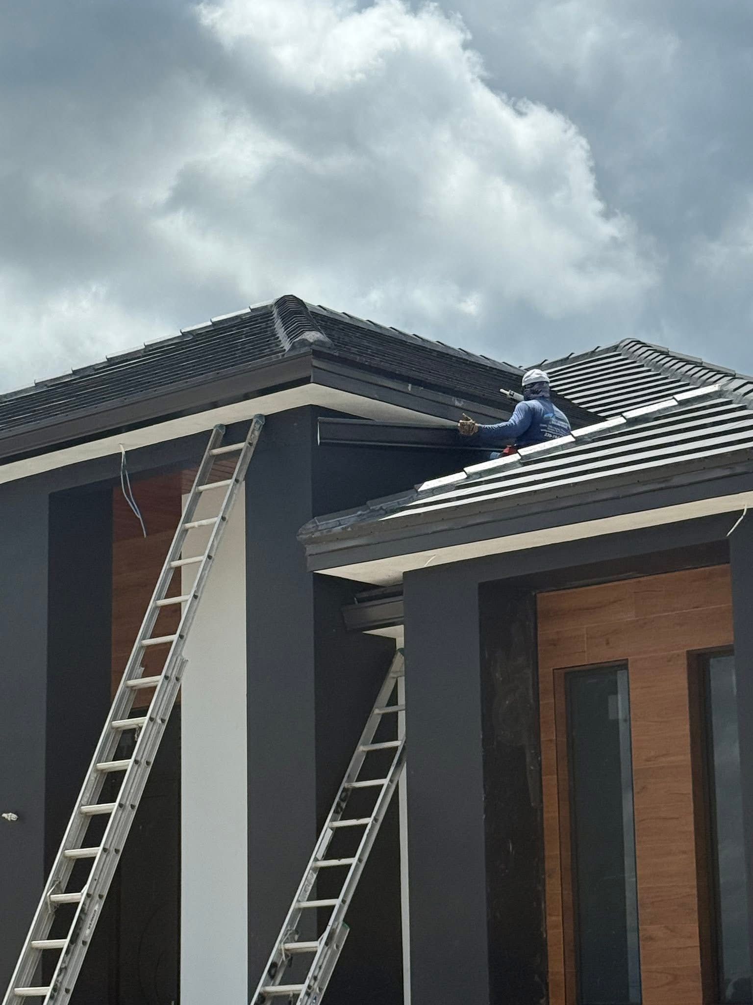 Worker on a ladder repairing a gray-tiled roof on a modern house