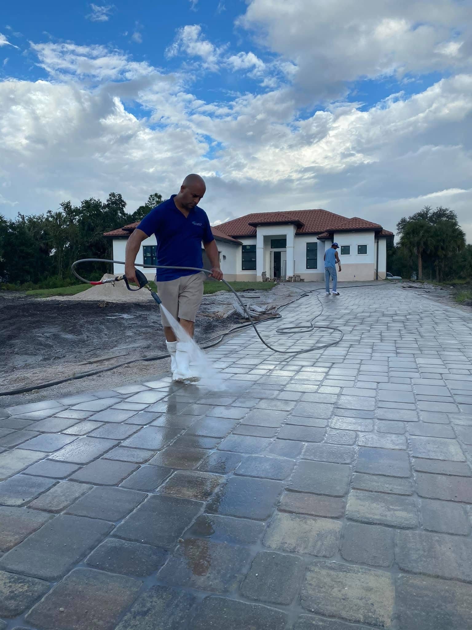 Two people cleaning a wet stone courtyard in front of a small house under a cloudy sky