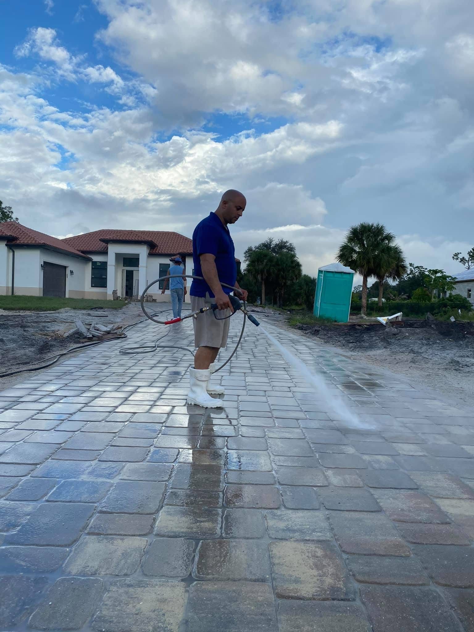 Person spraying water on a paved path beside houses under a cloudy sky.