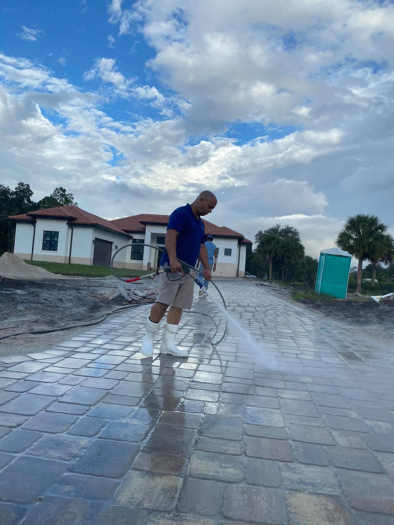 Person cleaning a wet cobblestone street with a hose under a cloudy sky near houses