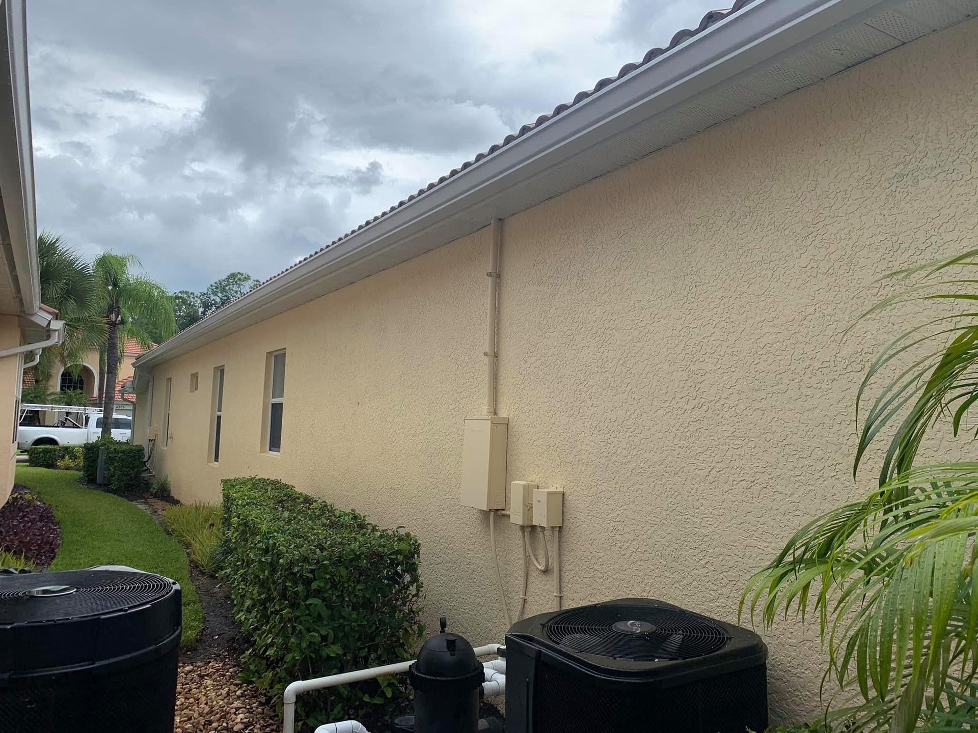 Side view of beige stucco house with roof overhang, utility boxes, and cloudy sky above a small lawn.
