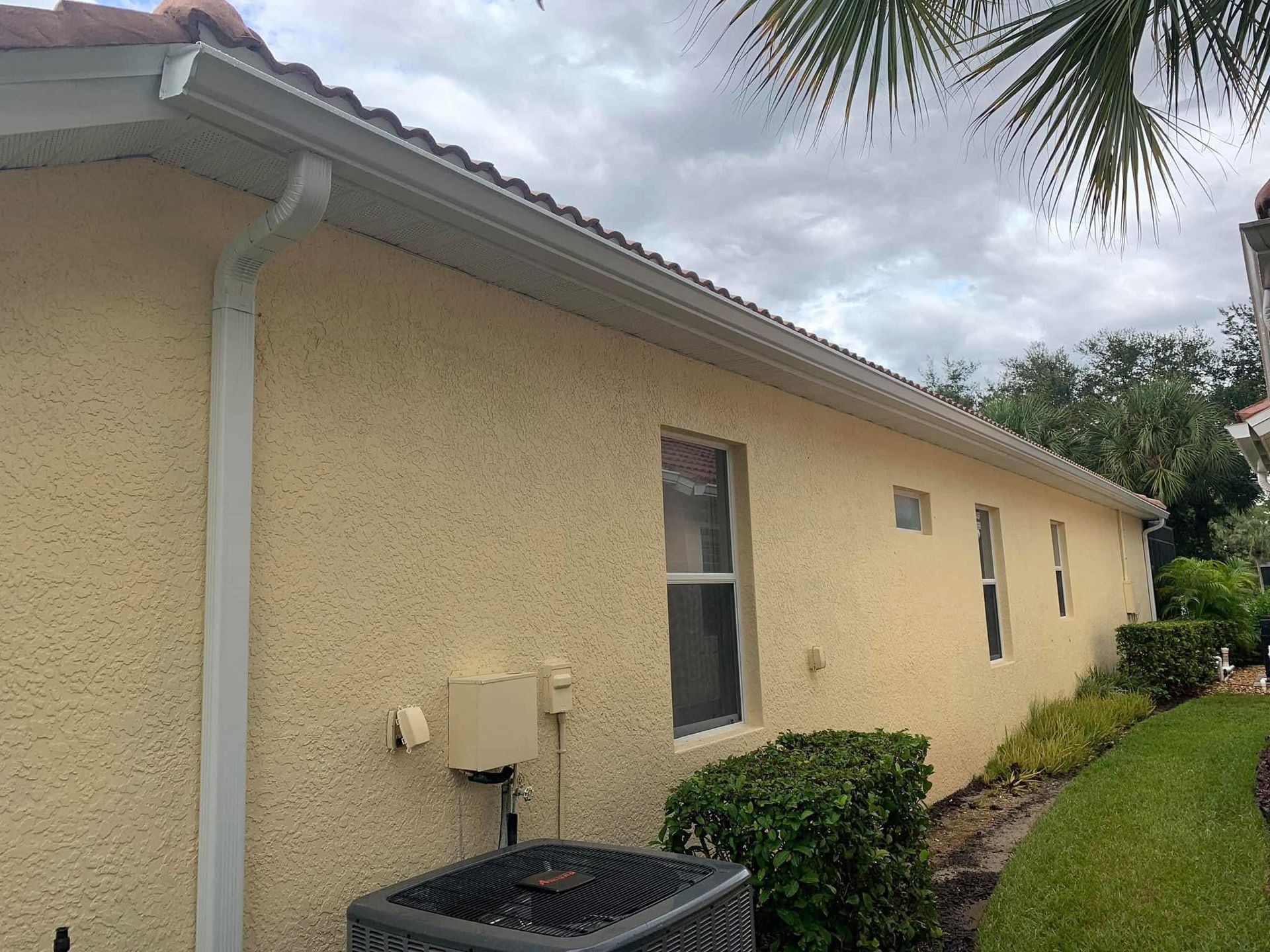 Side view of a beige stucco house with a gray roof, window, downspout, and neatly trimmed shrubs along the wall