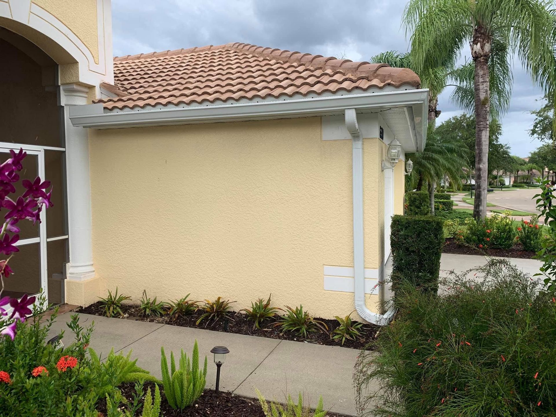 Yellow stucco house wall with terracotta roof beside a walkway and flowering garden.