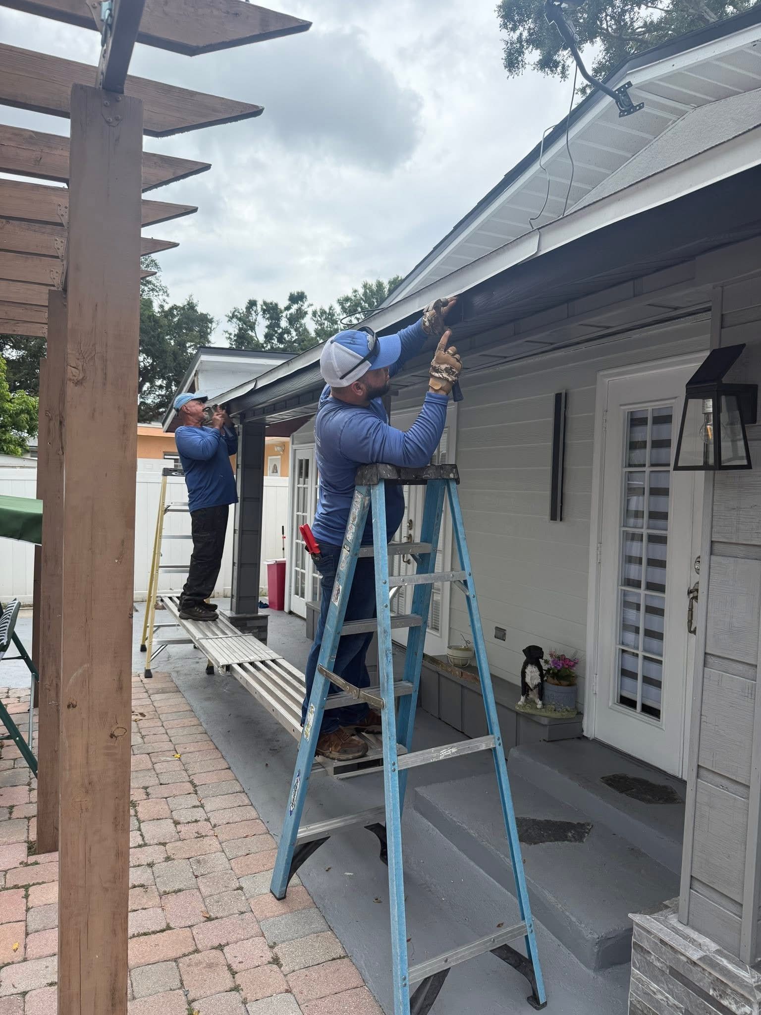 Workers on ladders repairing a white house’s roof and eaves on a porch.