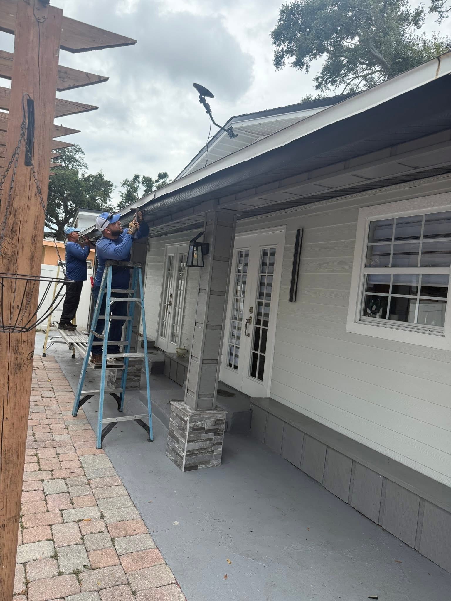 Workers on a ladder installing materials along the side of a white house with gray trim