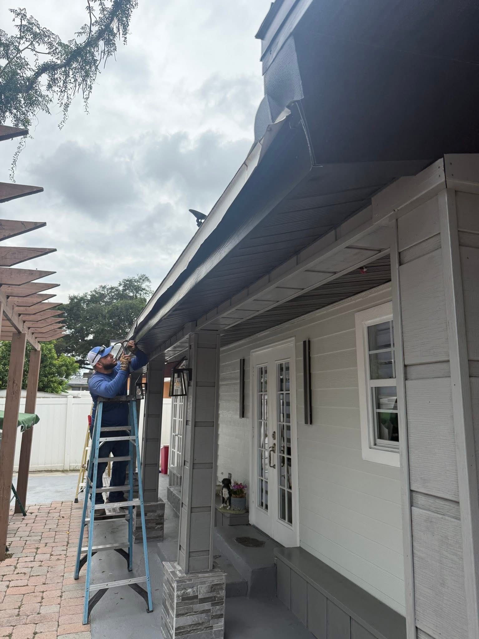 Worker on a ladder cleaning a house’s blue roof gutter beside a white exterior wall