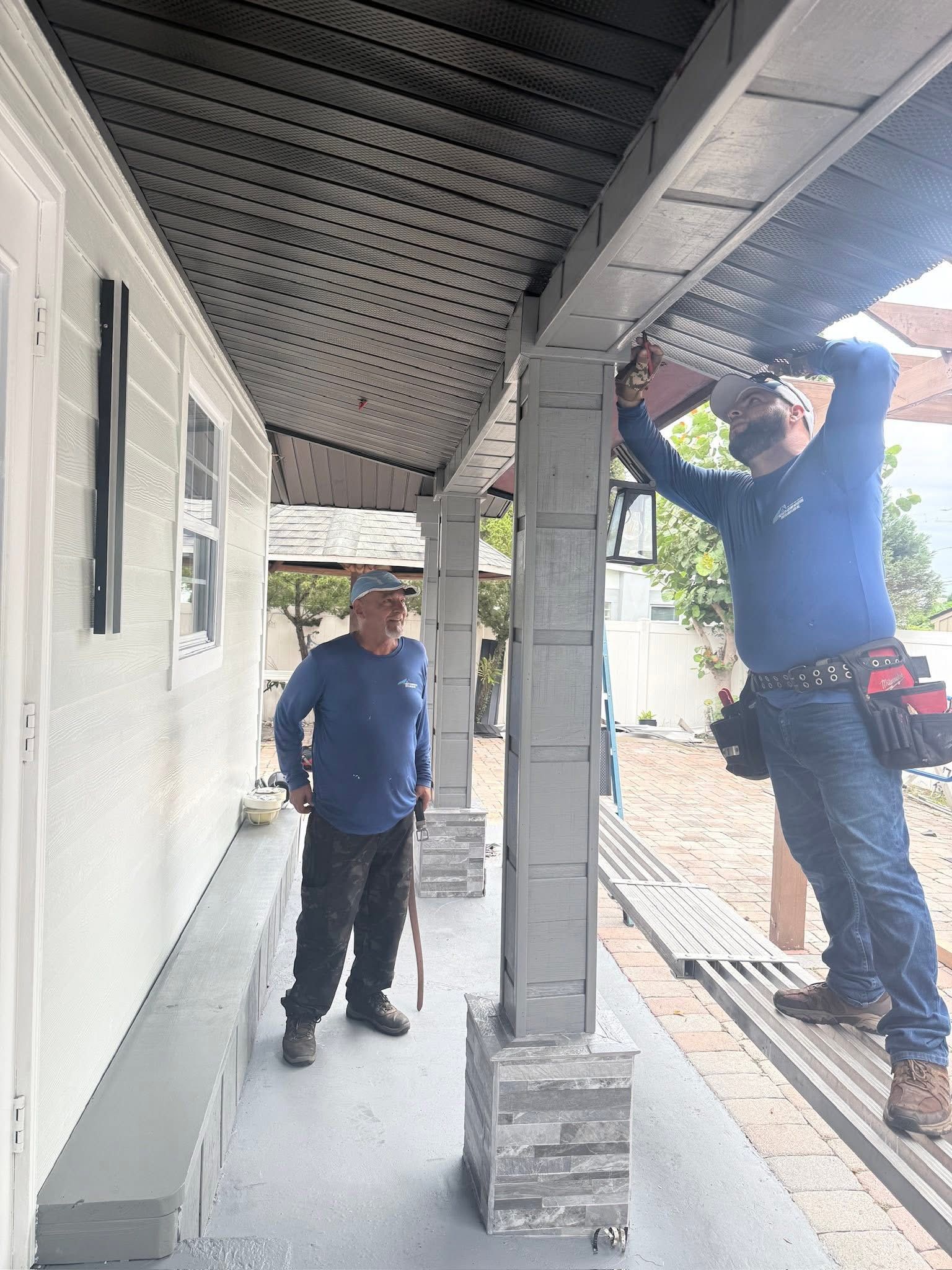 Two workers in blue shirts repair the underside of a covered walkway, one standing on a ladder.