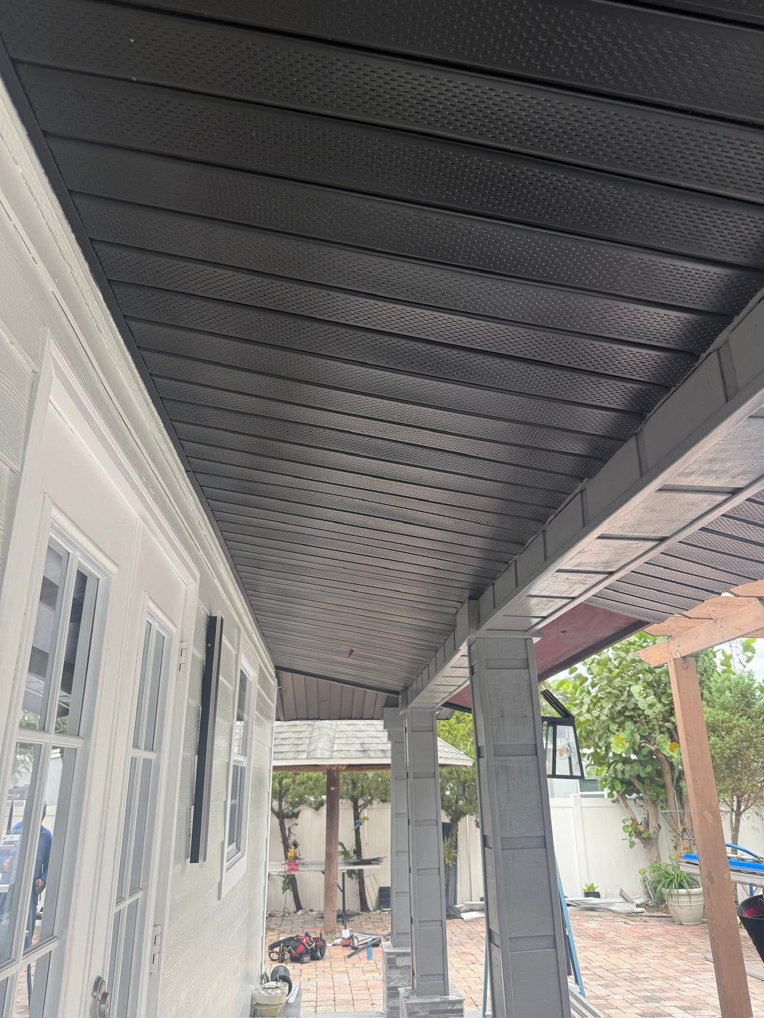 Covered backyard patio with a dark perforated ceiling, white siding, and construction materials on the ground.