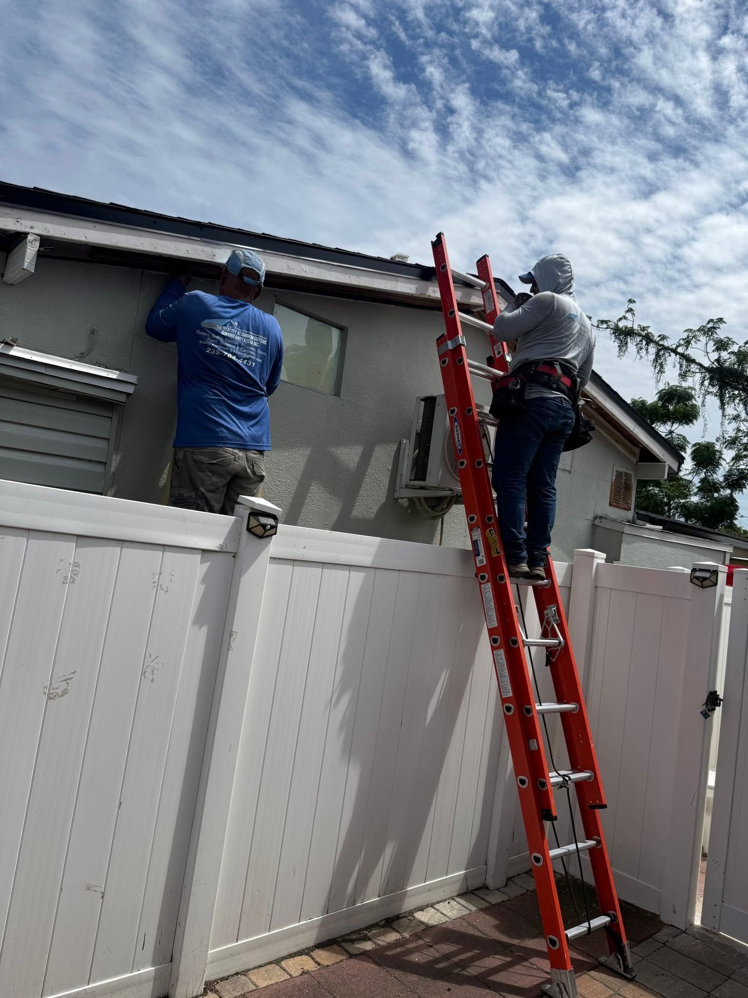 Two workers on a ladder repairing a roof above a white fence.