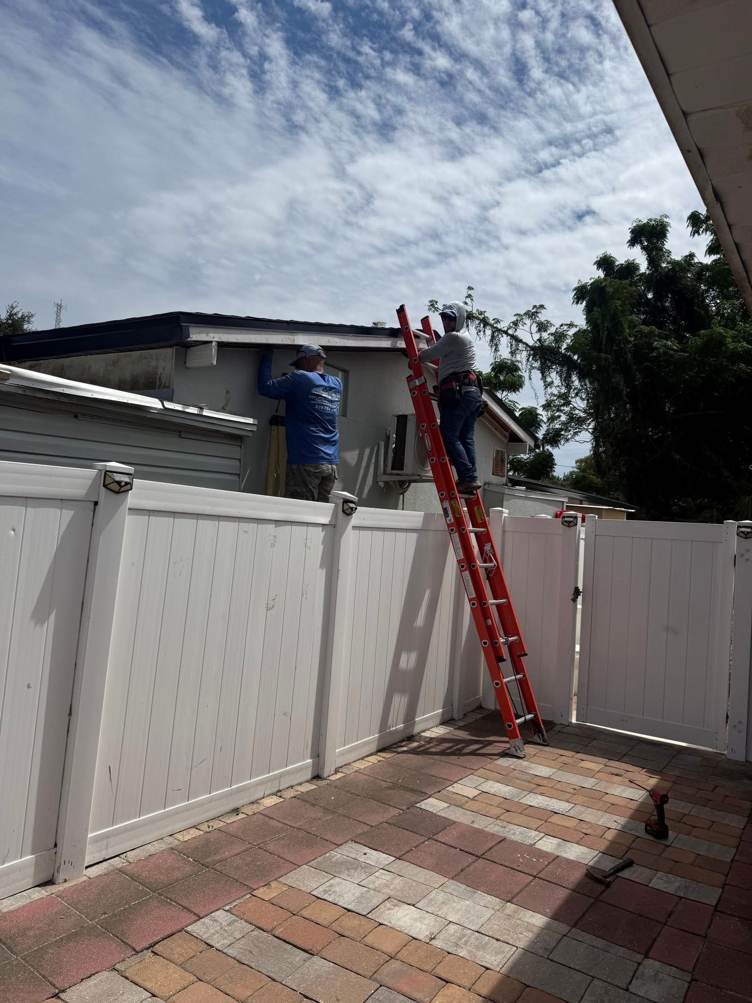 Two workers on ladders fixing a roof behind a white fence in a sunny backyard