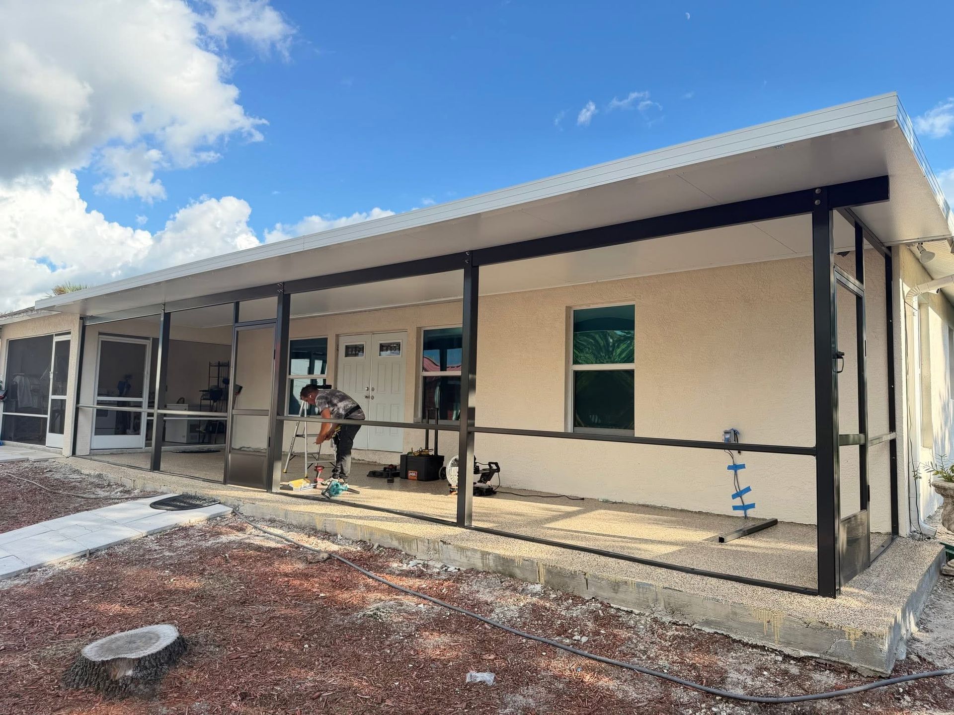 Single-story building under construction with workers, beige walls, and a covered porch under a blue sky