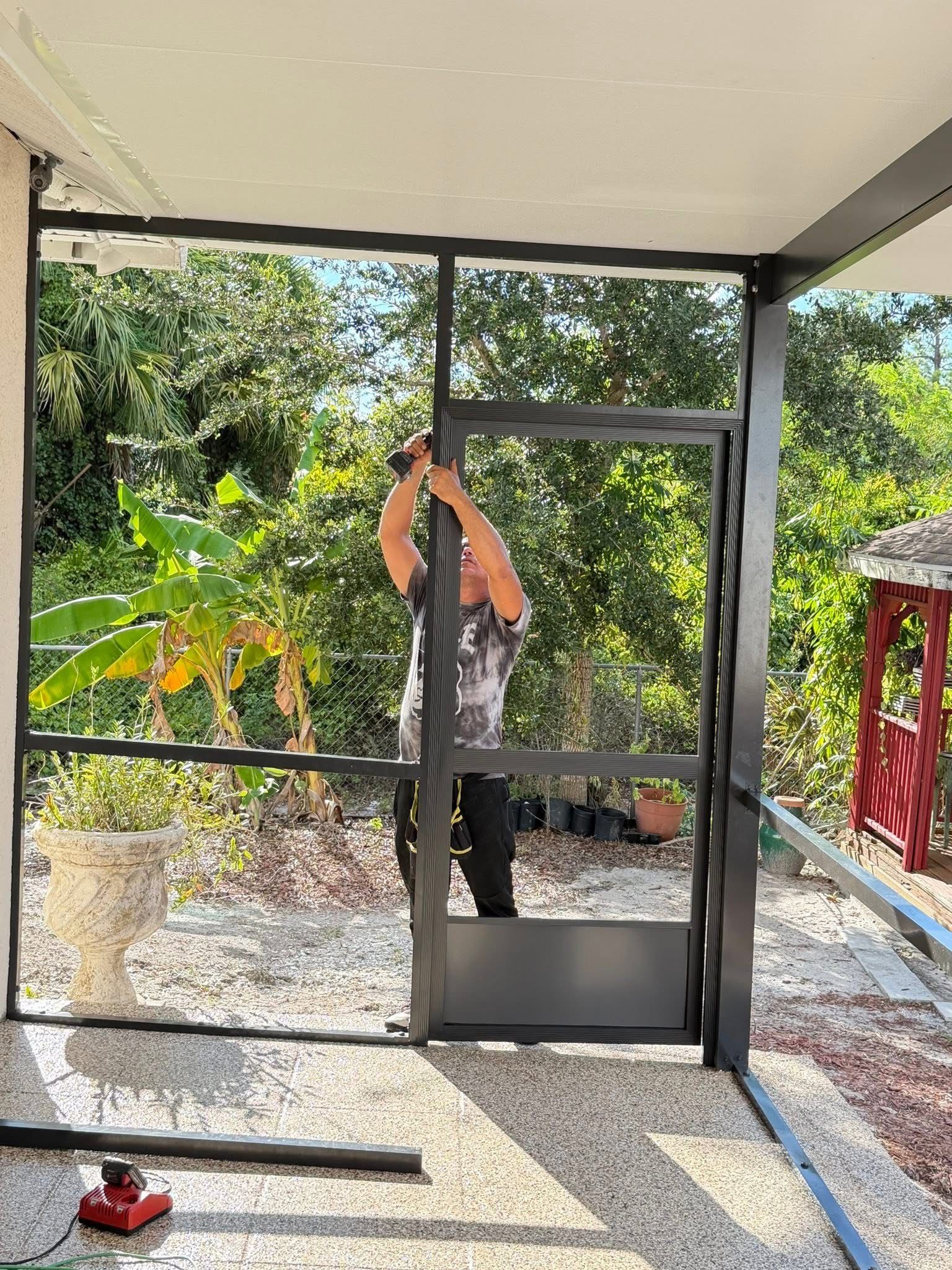 Person installing a black-framed screen door on a porch with trees and gravel outside