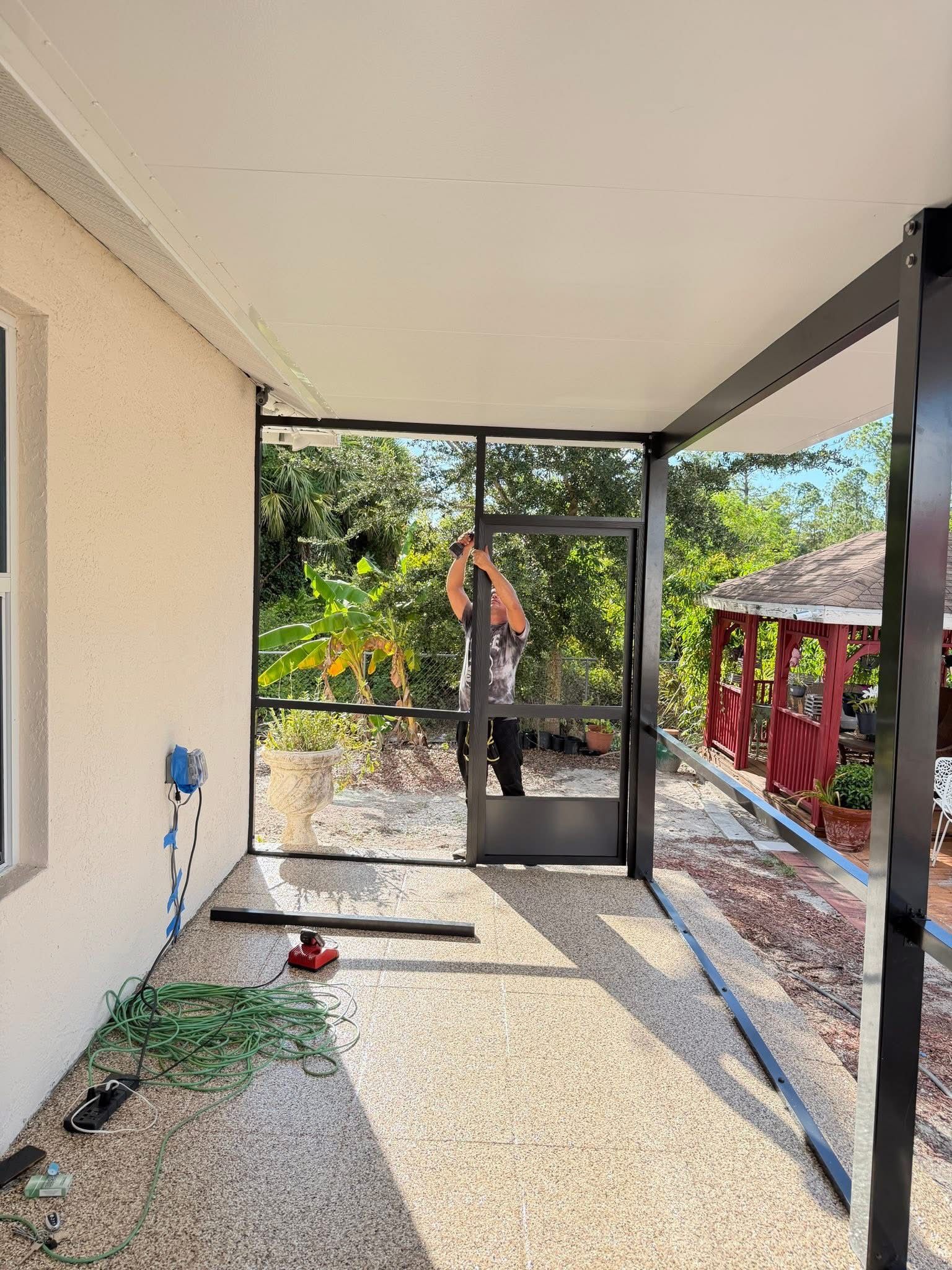Covered patio with a person standing by a black metal gate, overlooking a yard and garden area.