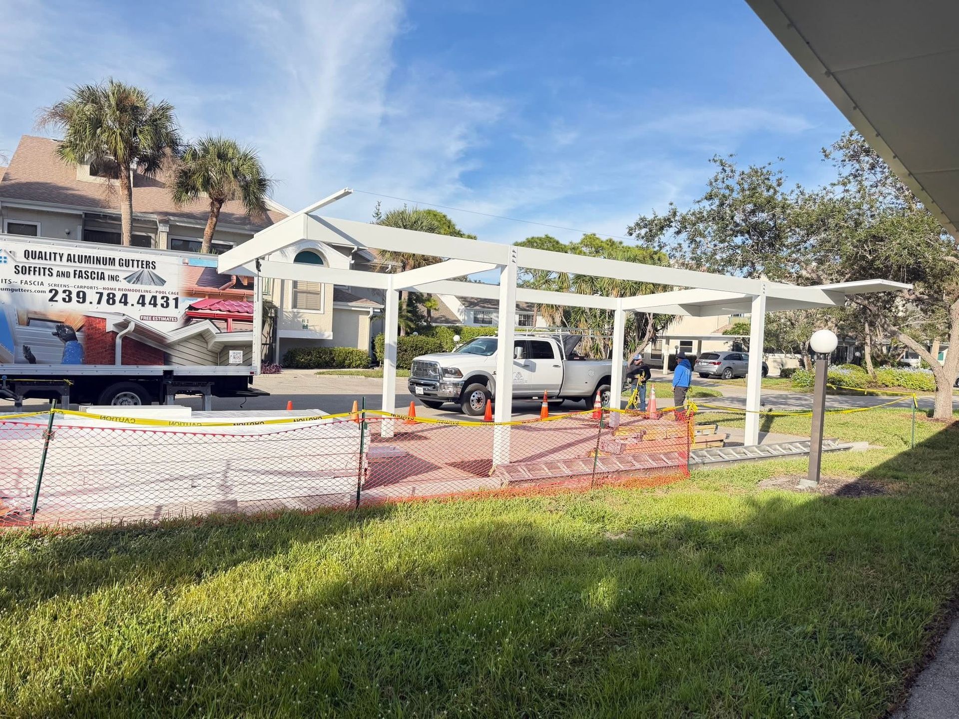 White canopy structure over a grassy area with parked cars and orange construction fencing on a sunny day