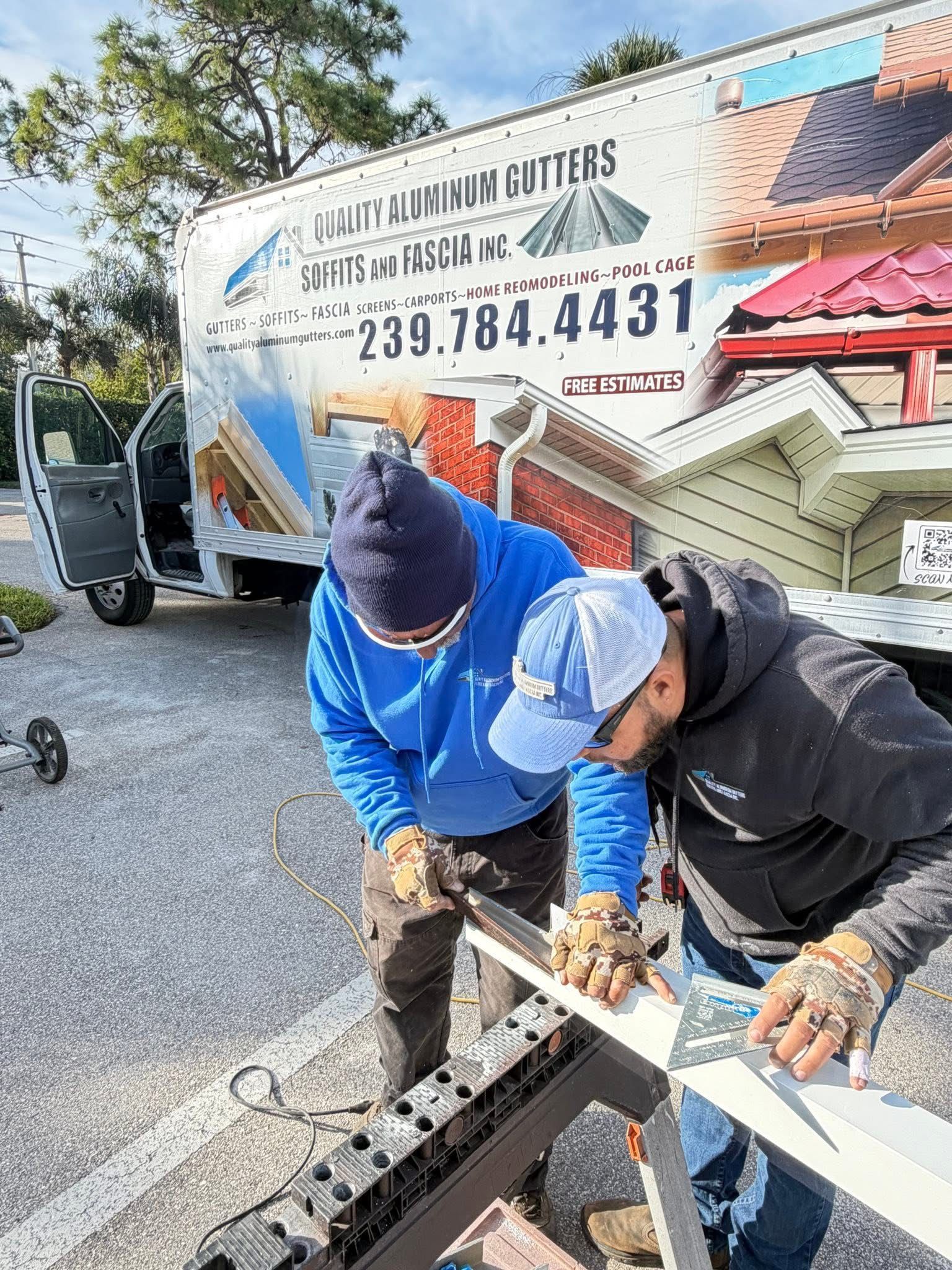 Two workers in blue and black crouch over a metal part beside a utility truck.