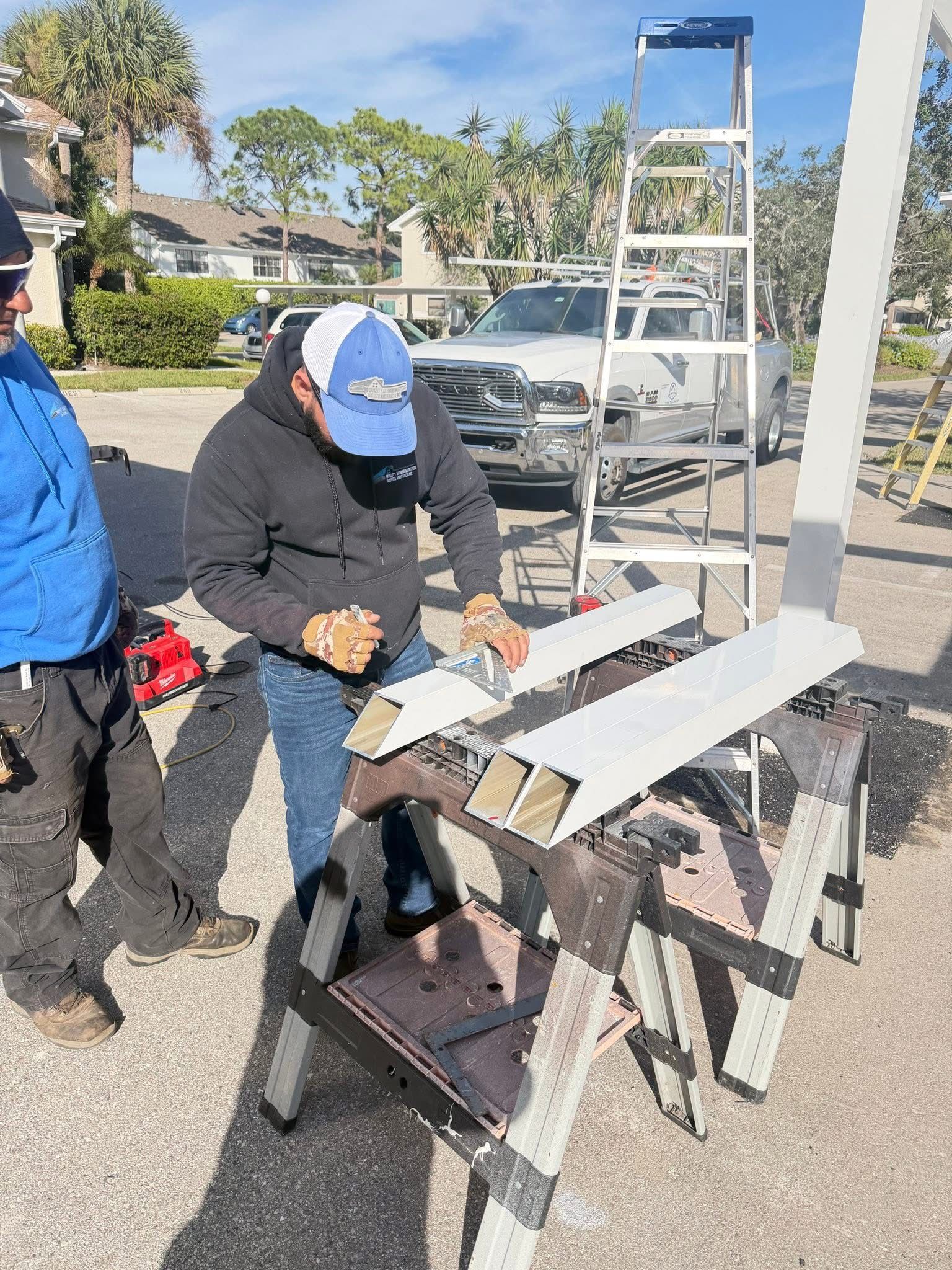 Two workers assembling a metal frame outdoors beside a ladder and a parked car.