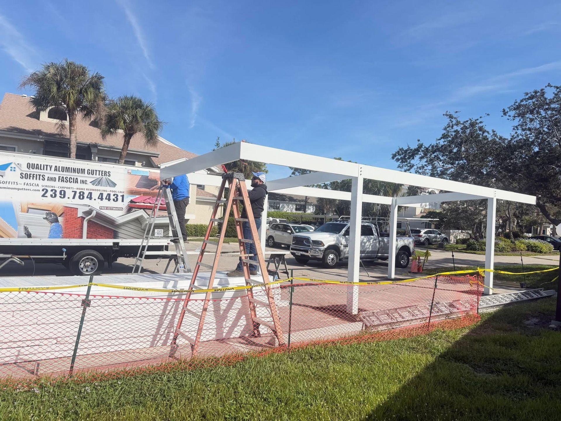 White canopy tent frame over a paved area, with a ladder and parked vehicles in the background.
