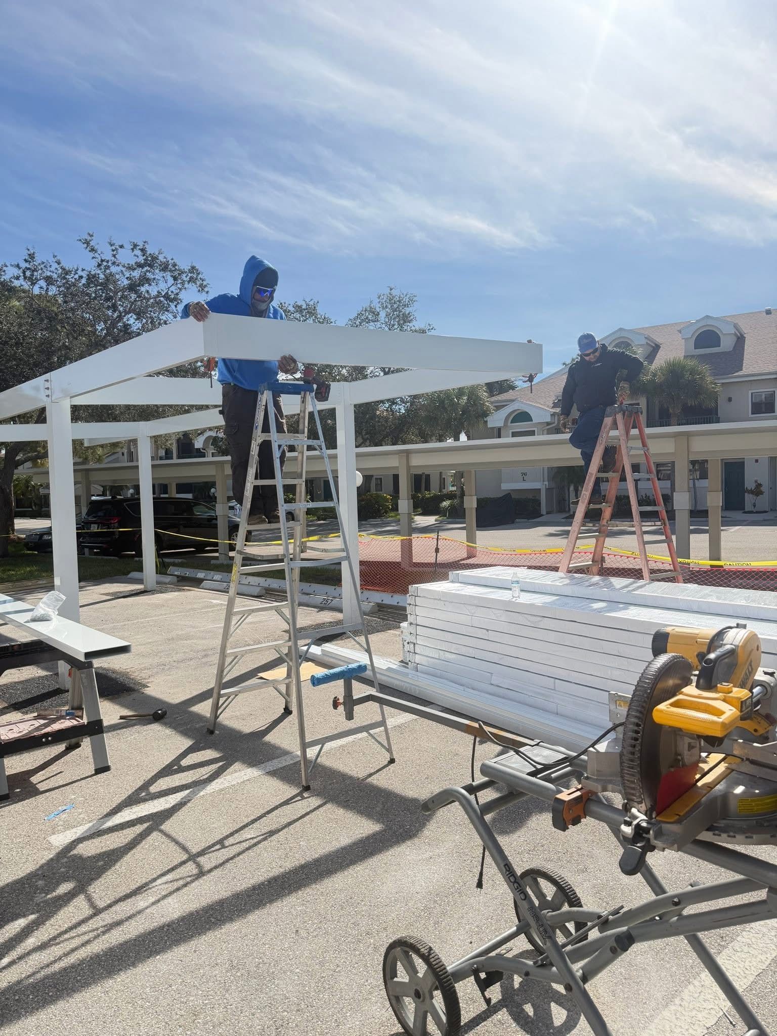 Worker on ladder assembling a white pergola in an outdoor worksite with tools and materials nearby