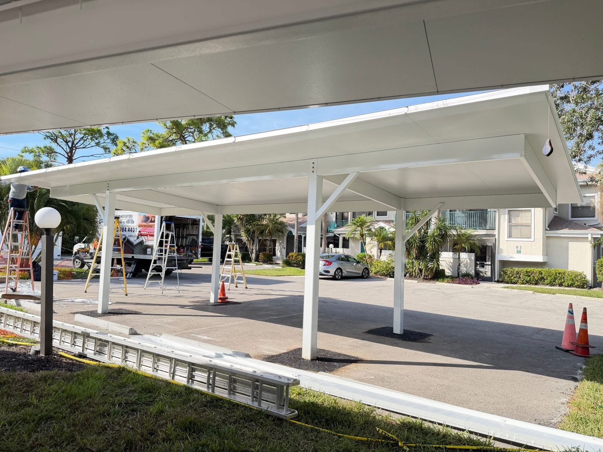 Covered parking area with white steel canopy and several orange traffic cones in a lot outdoors