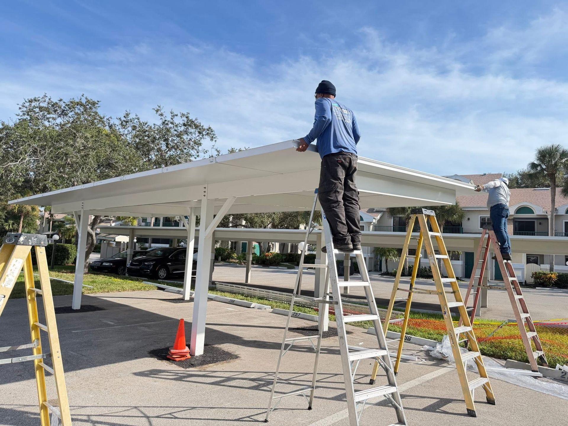 Two workers installing a white metal carport roof, with ladders and traffic cones in a driveway.