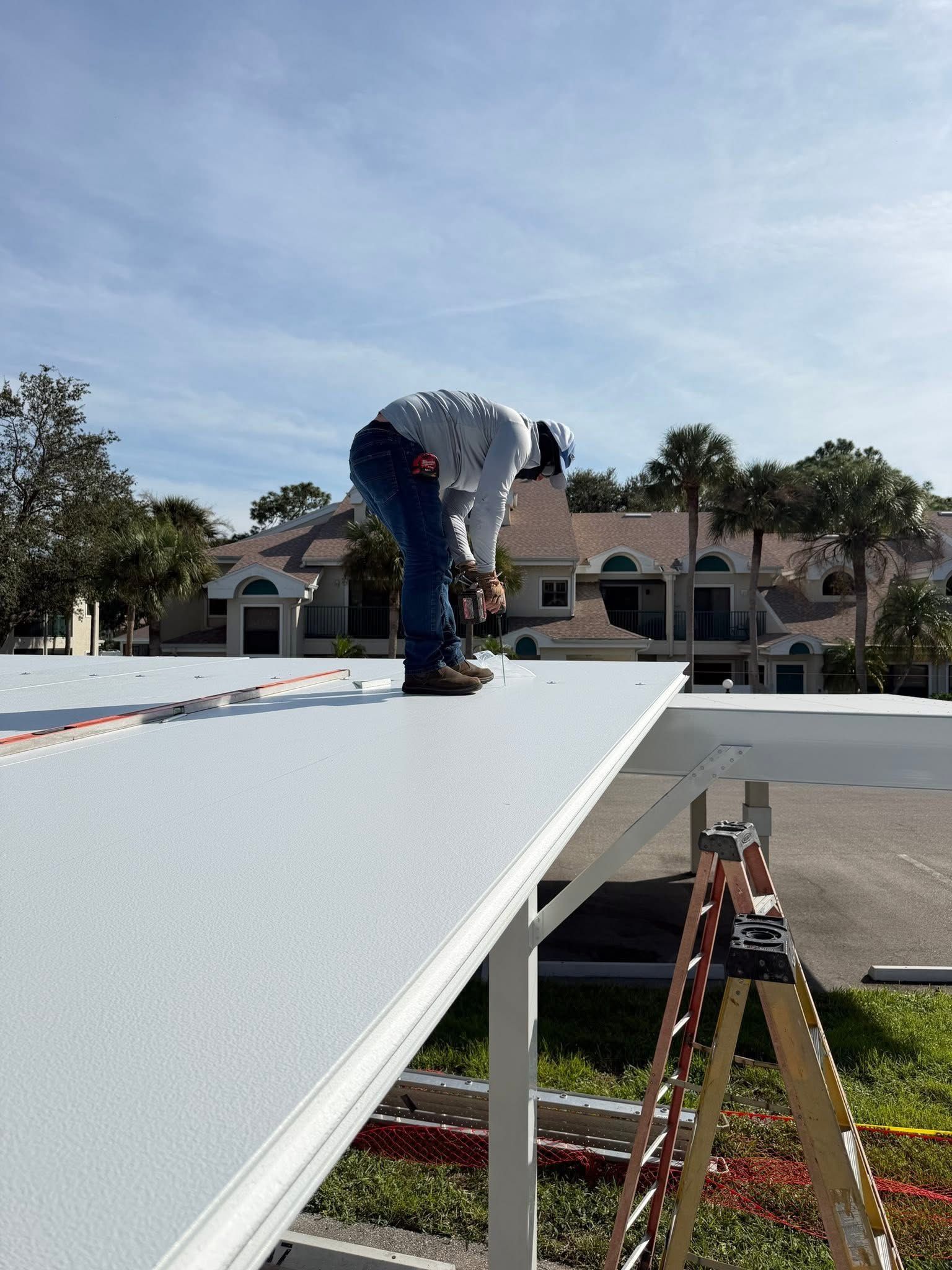 Worker installing a white roof panel on a building, with a ladder and suburban houses in the background.