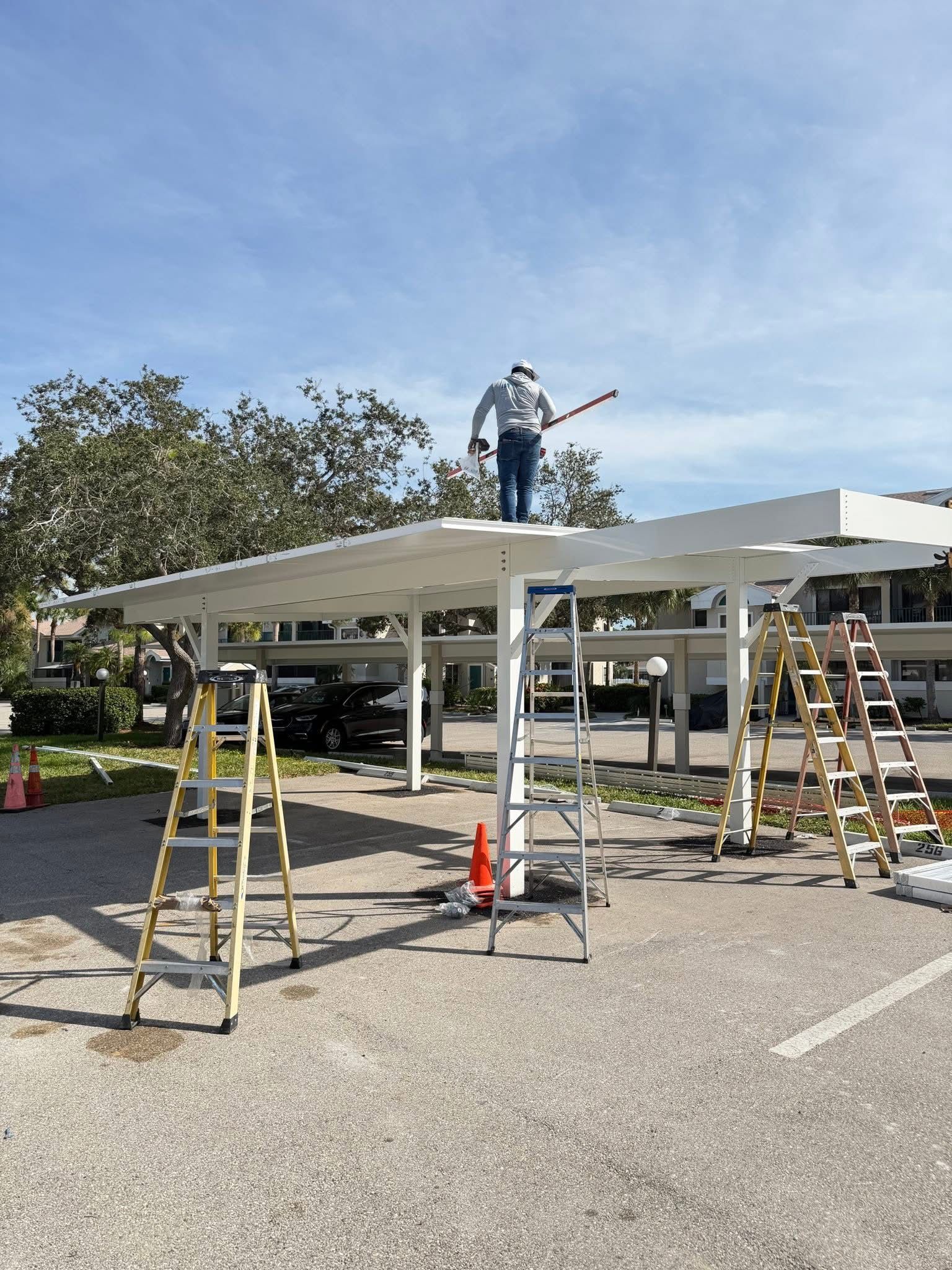 Worker painting a white canopy roof outdoors, with ladders and tools on a parking lot.
