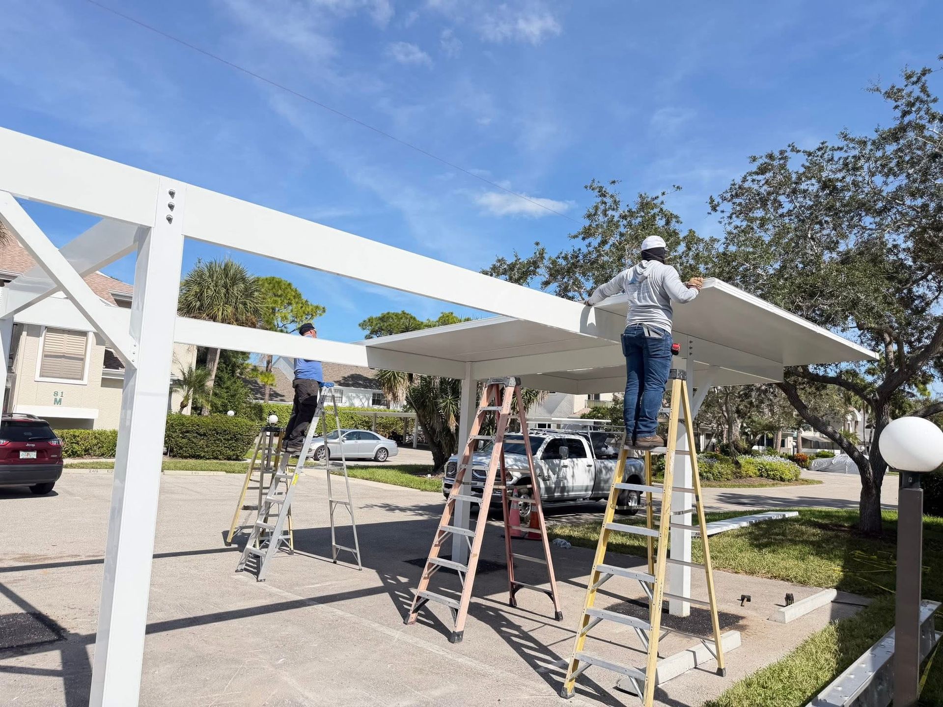 Workers installing a white canopy frame in a sunny driveway, with ladders and a parked car nearby