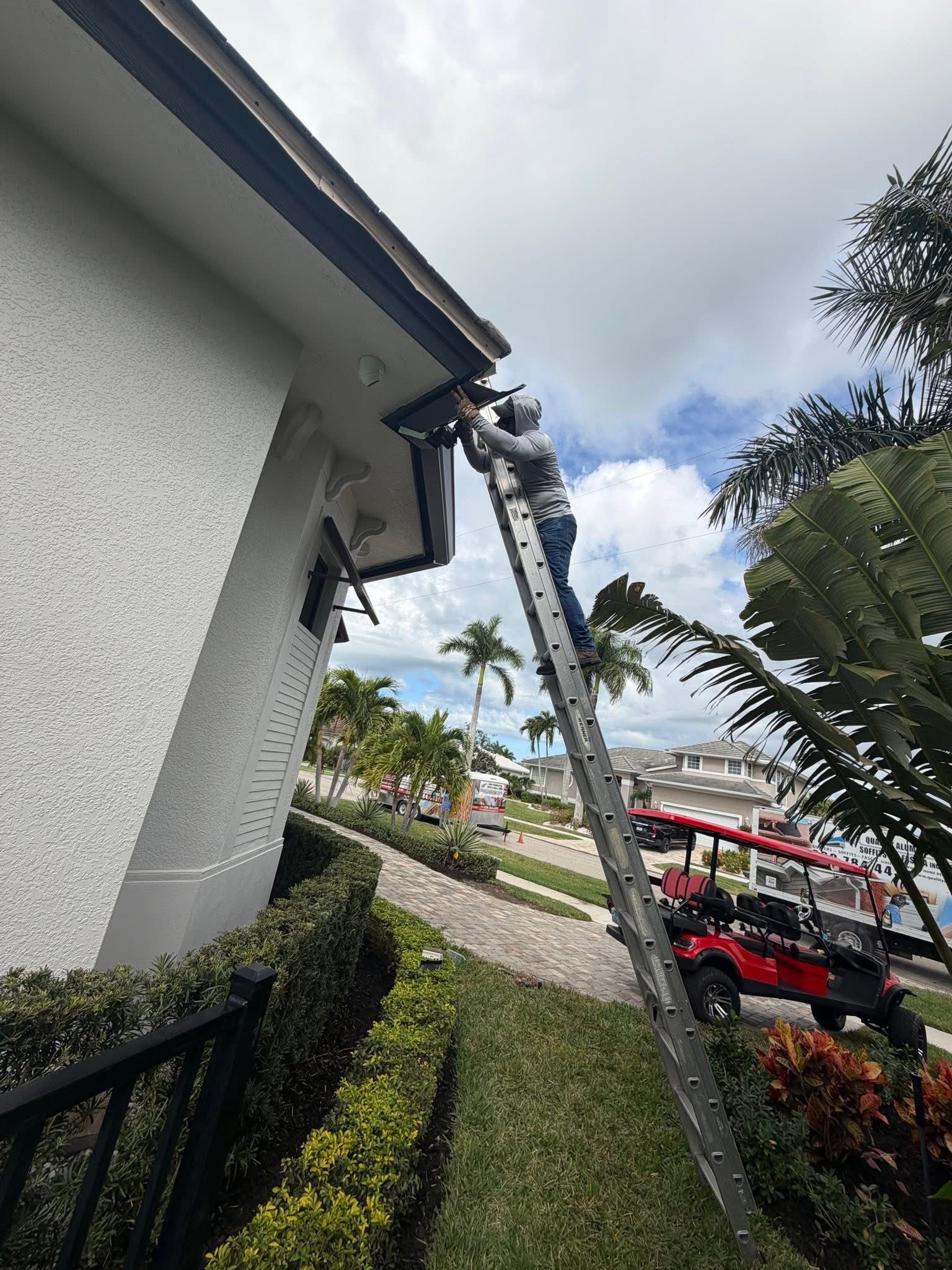 Ladder leaning against house roof edge beside a white wall and garden, with a red vehicle in the background