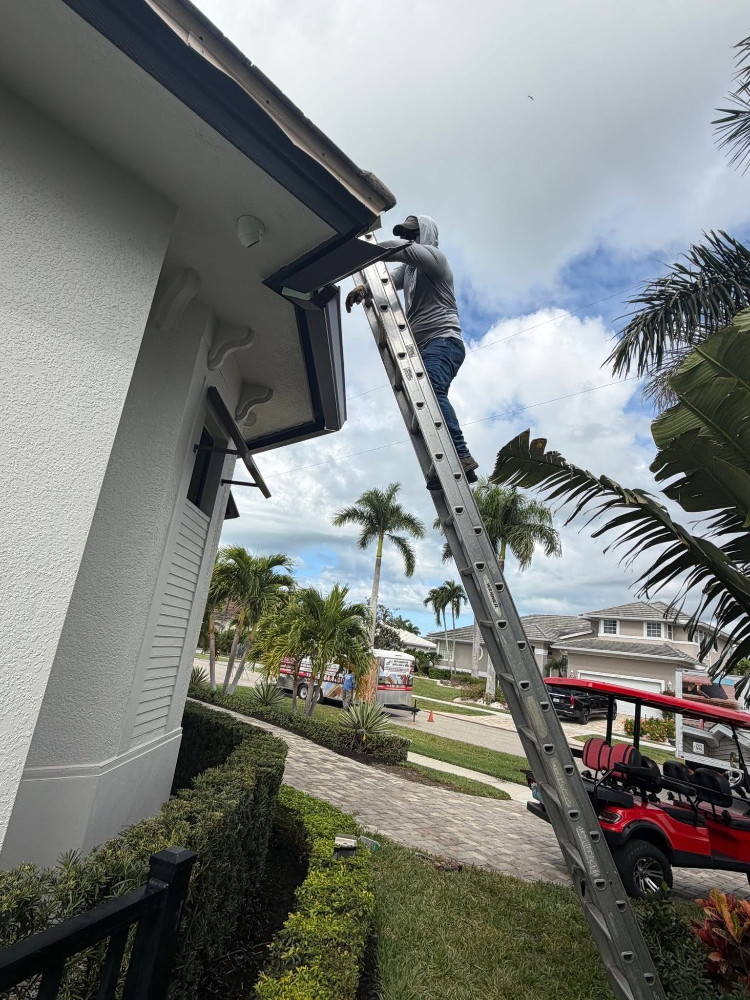 Worker on a ladder cleaning a house gutter beside a landscaped yard and parked red cart in the background