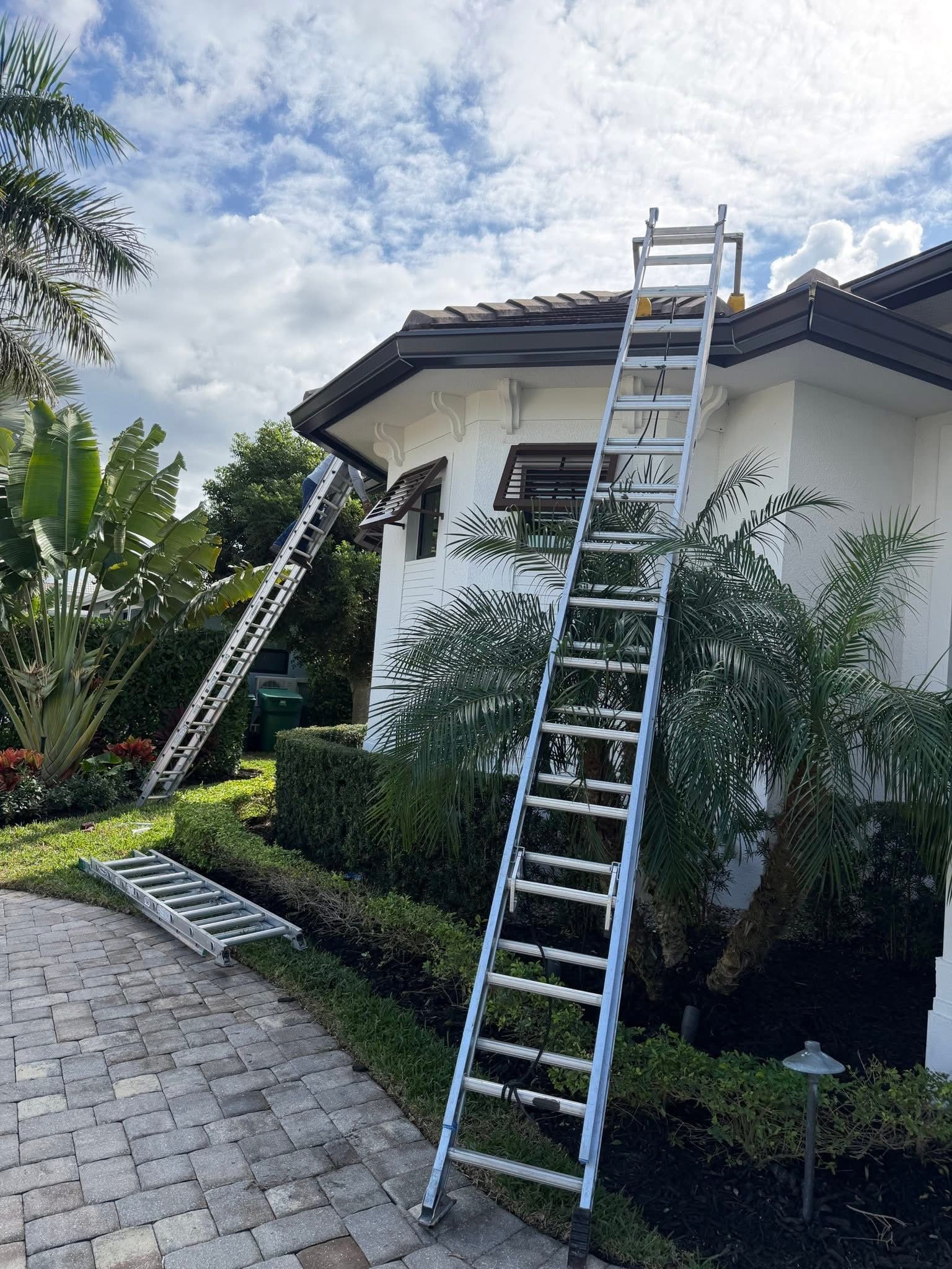 Two ladders leaned against a white house with a tiled roof and garden path under a cloudy sky