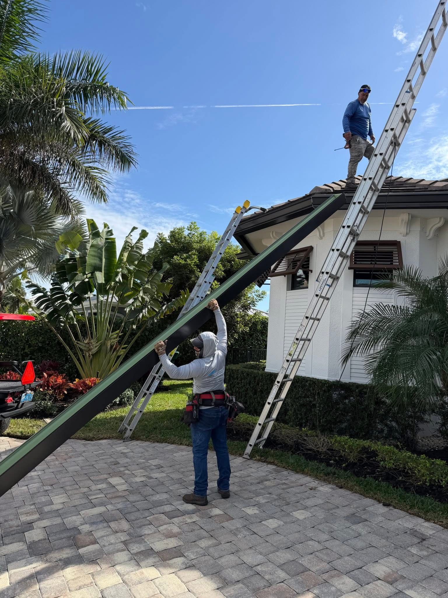 Two workers on ladders painting a white house roof under a blue sky.