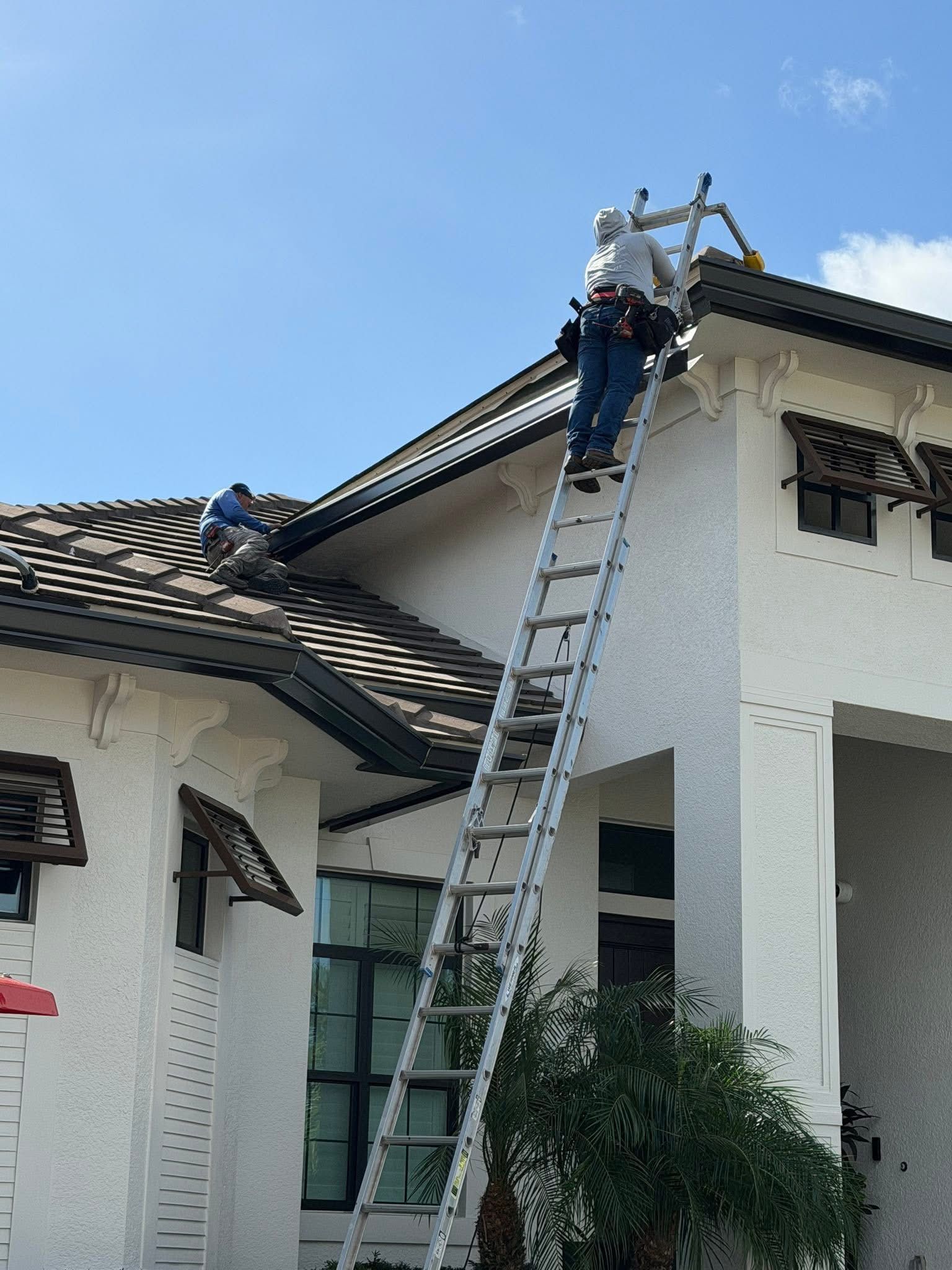 Workers on a tall ladder repairing the roof and gutter of a white house