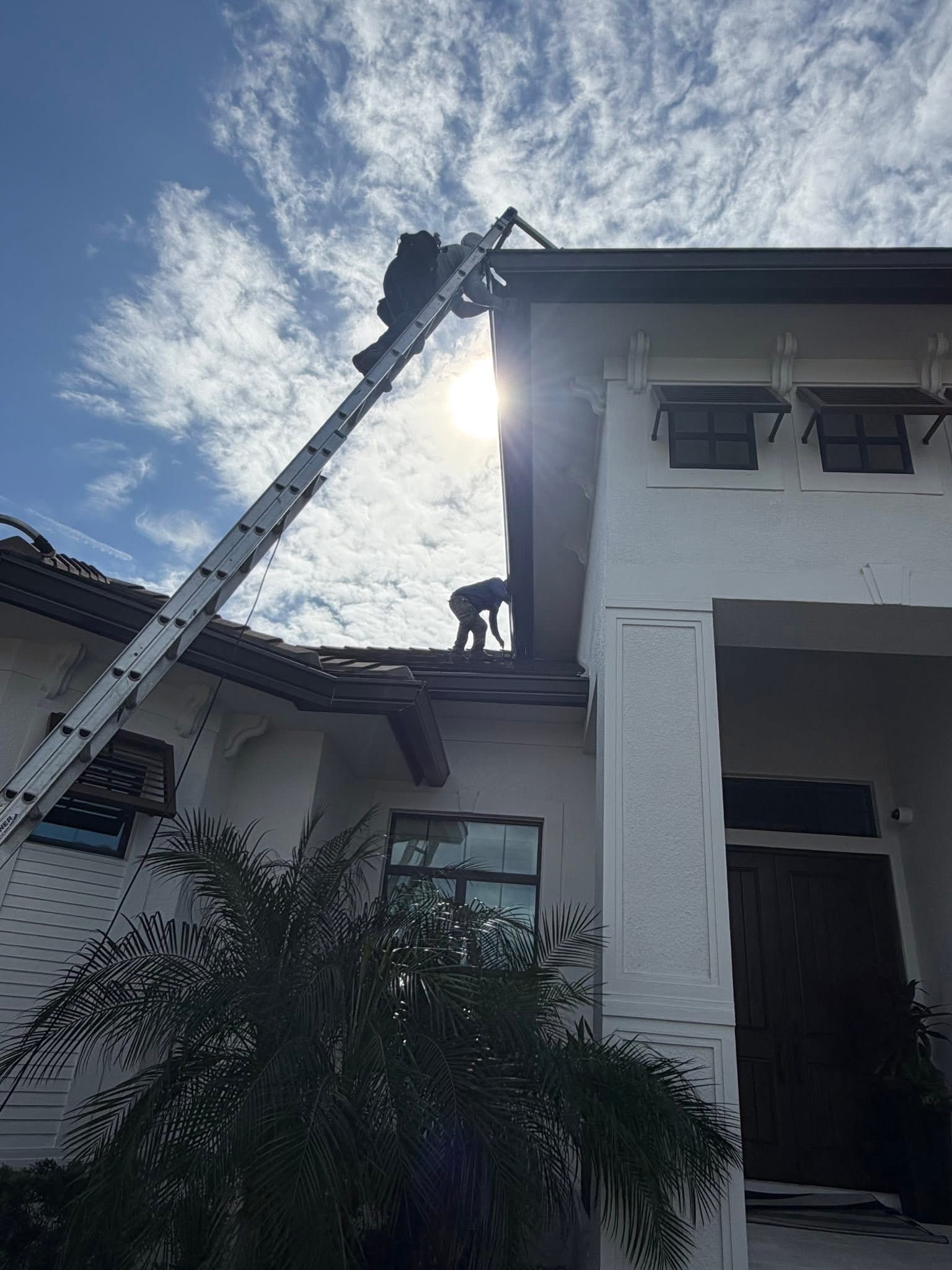 Person on a ladder near a house roof, with sunlight behind and cloudy sky above