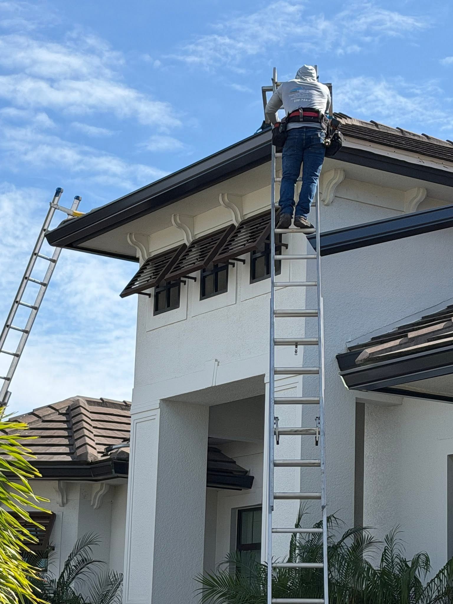 Worker on a ladder repairing the roof of a white house with a second ladder nearby.