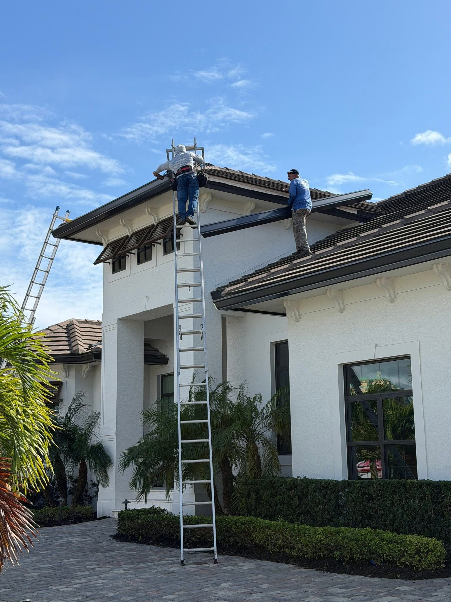 Workers on a ladder repairing the roof of a white house under a blue sky.