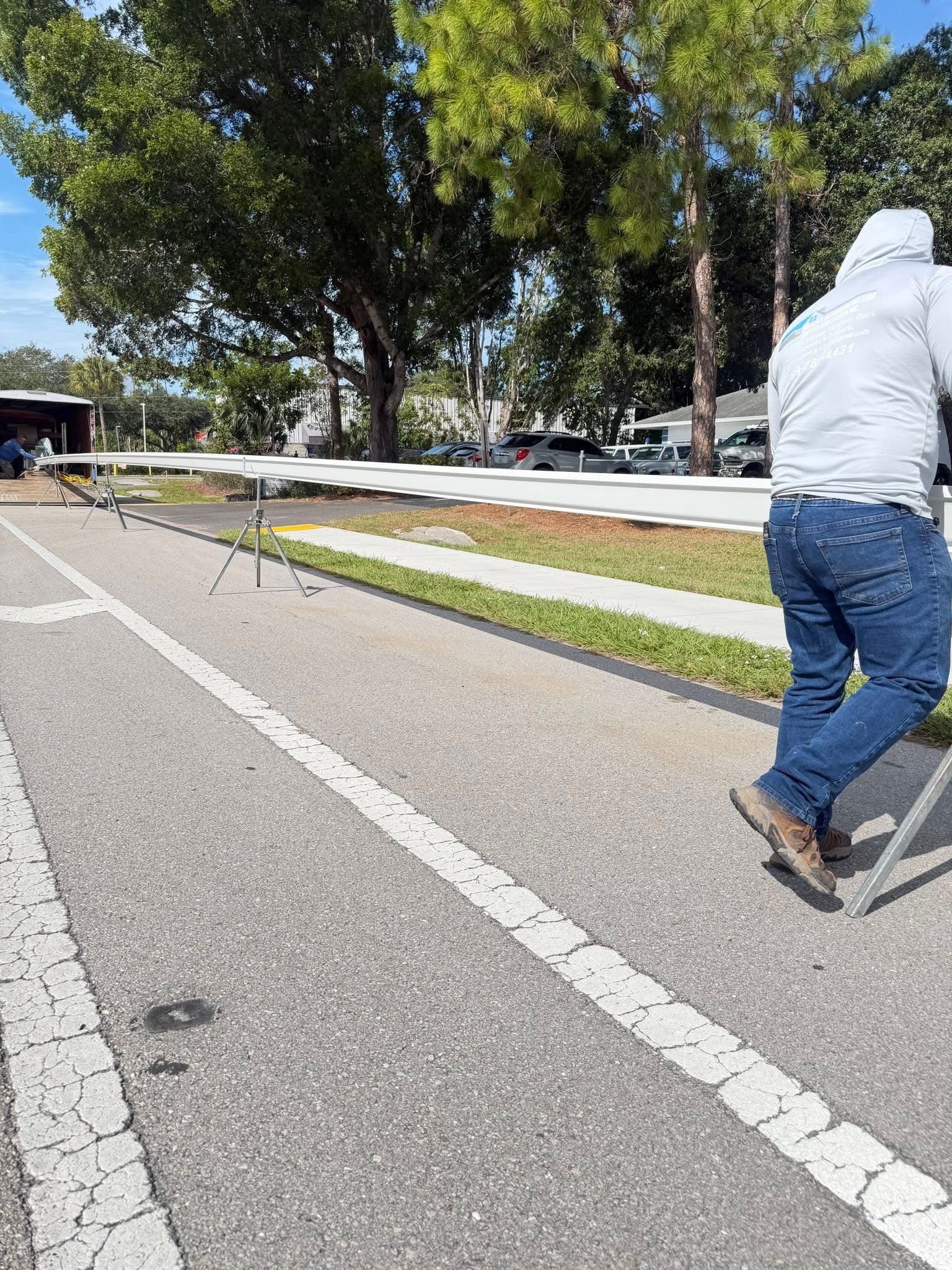 Person in a gray hoodie and jeans pushing a stroller along a road beside trees and a grassy median.
