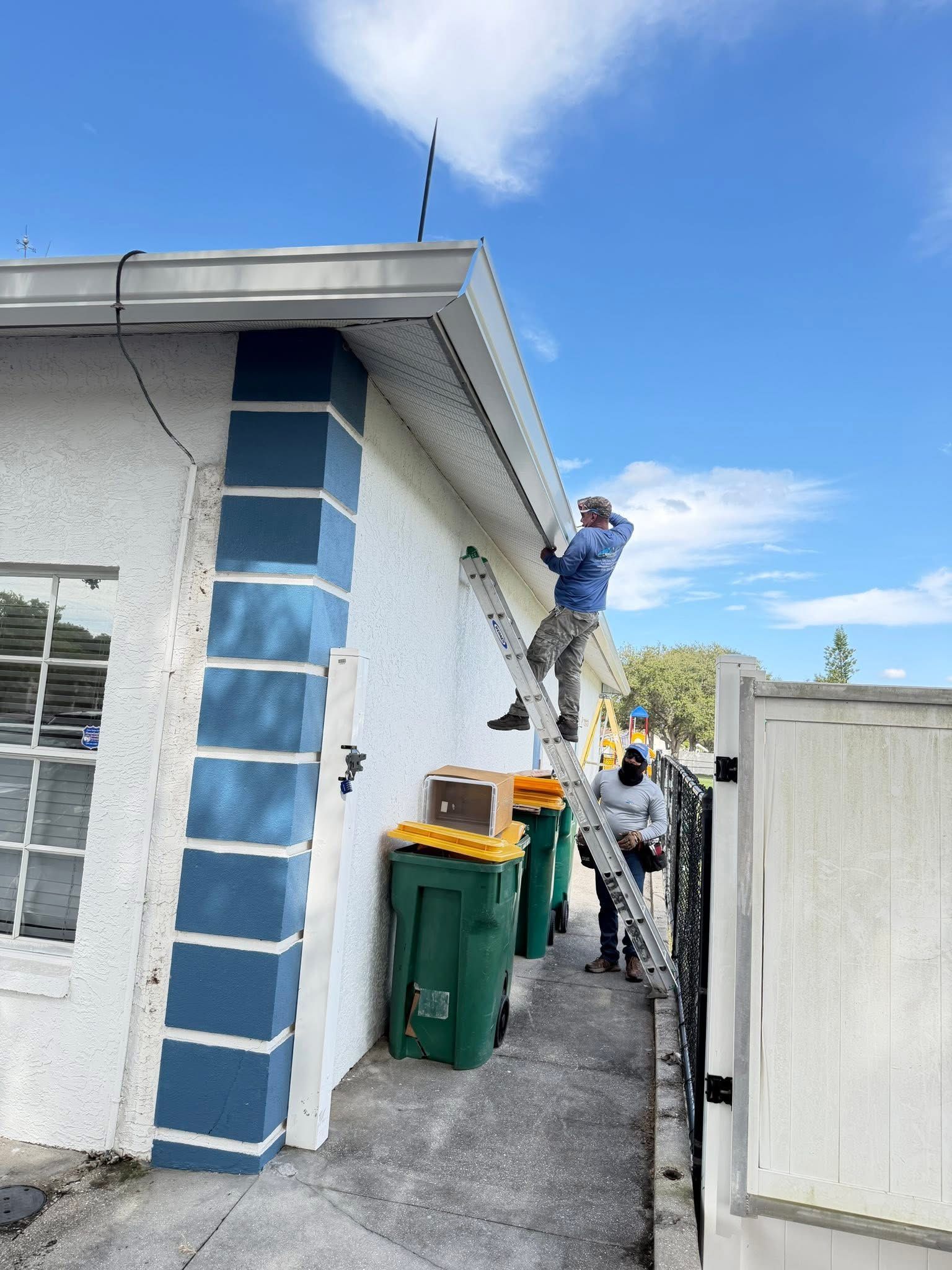 Worker on a ladder painting the side of a house beside trash bins and a white gate
