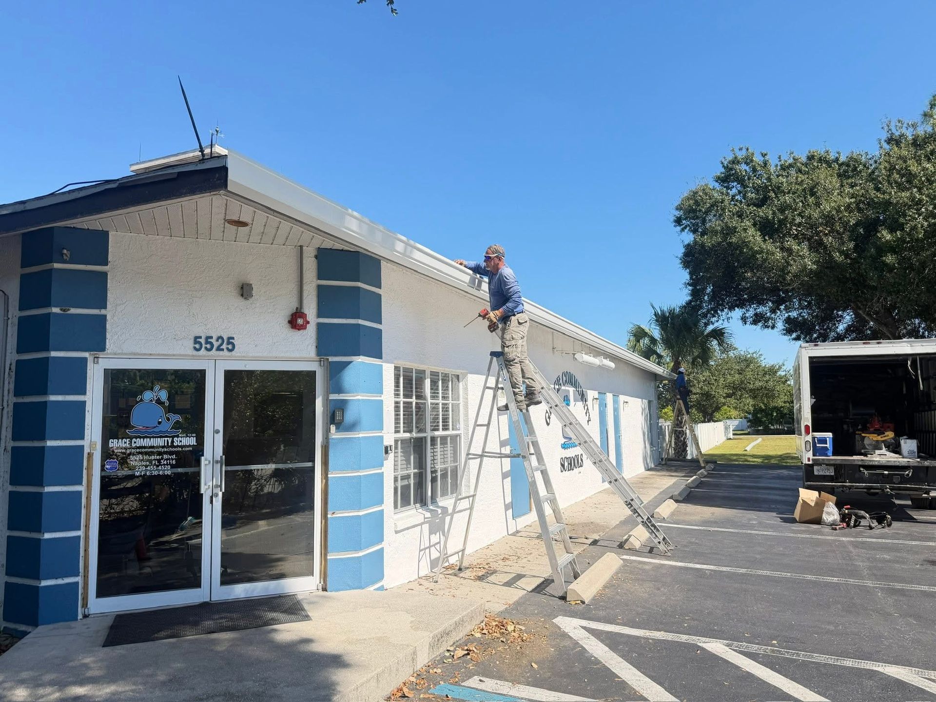 Two workers on ladders painting the exterior of a white building with blue trim on a sunny day