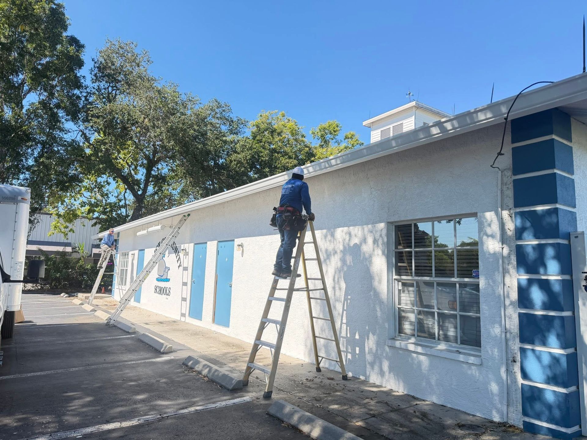 Worker on ladder painting the exterior of a white building with blue trim under a clear sky.