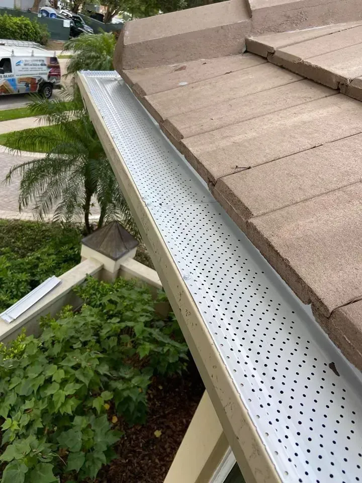 Close-up of a house roof gutter with white perforated leaf guard beside brown shingles.