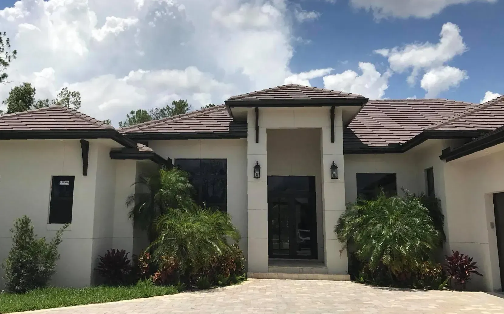 Modern white house with brown tile roof and front entrance framed by palm plants under a cloudy sky