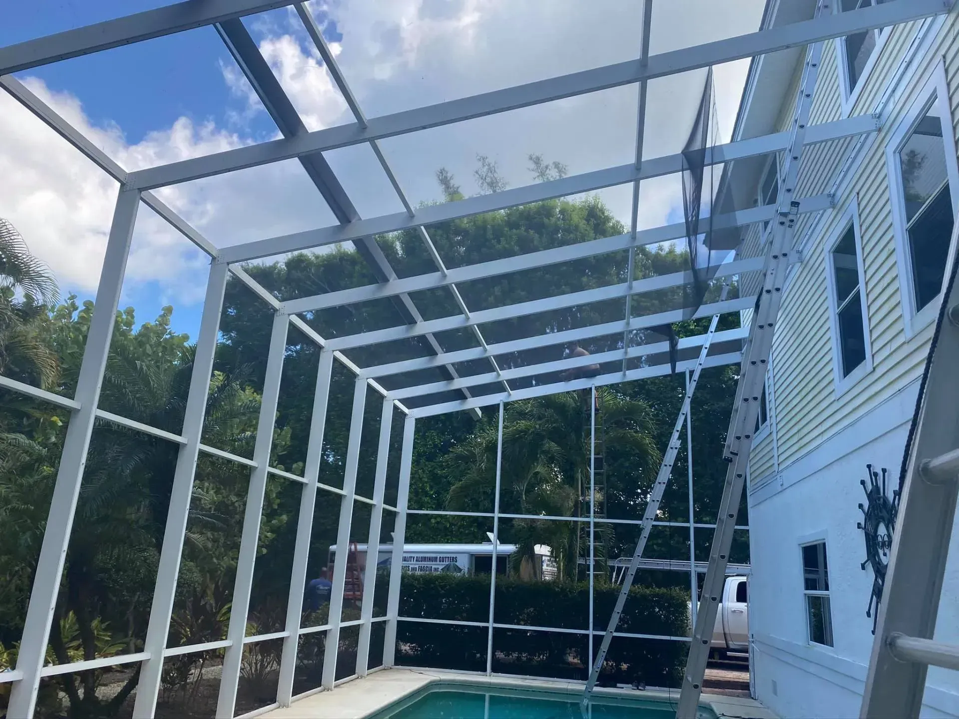 White pergola frame under construction beside a house, with a screened roof and blue sky overhead