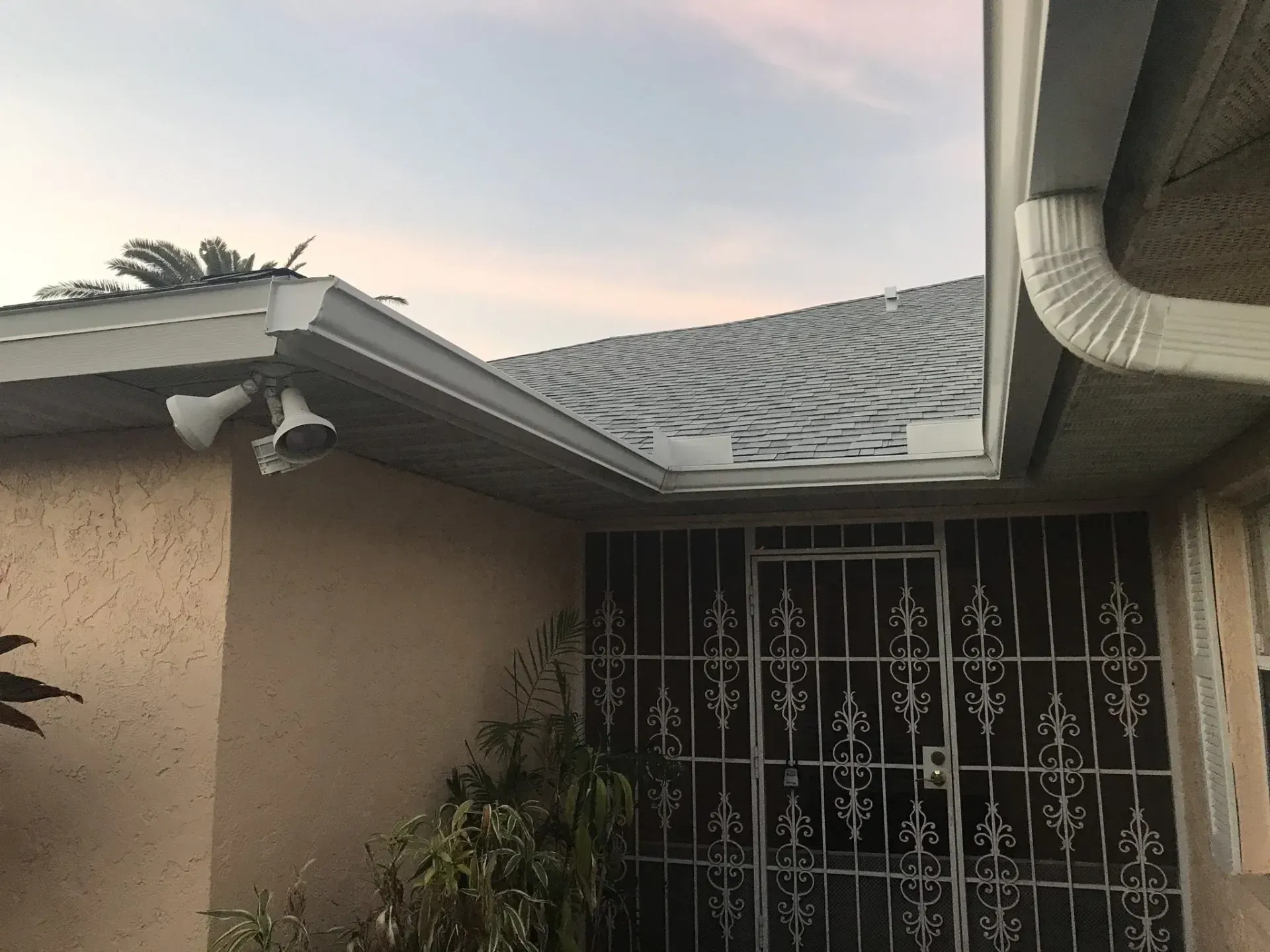 Suburban house exterior with beige stucco, gray roof, and a white metal security gate at dusk