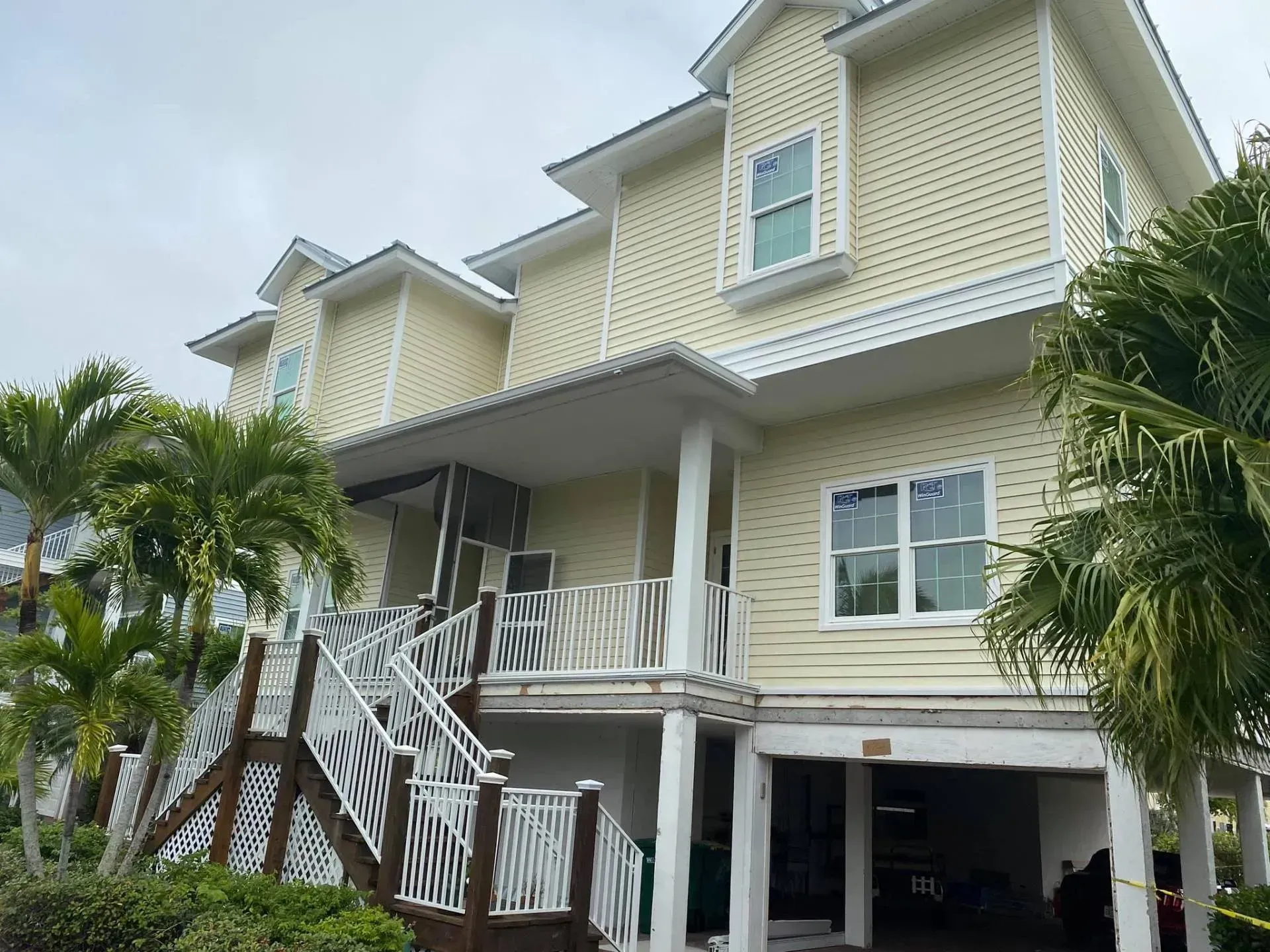 Raised beach house with white stairs, balconies, and palm trees under a cloudy sky