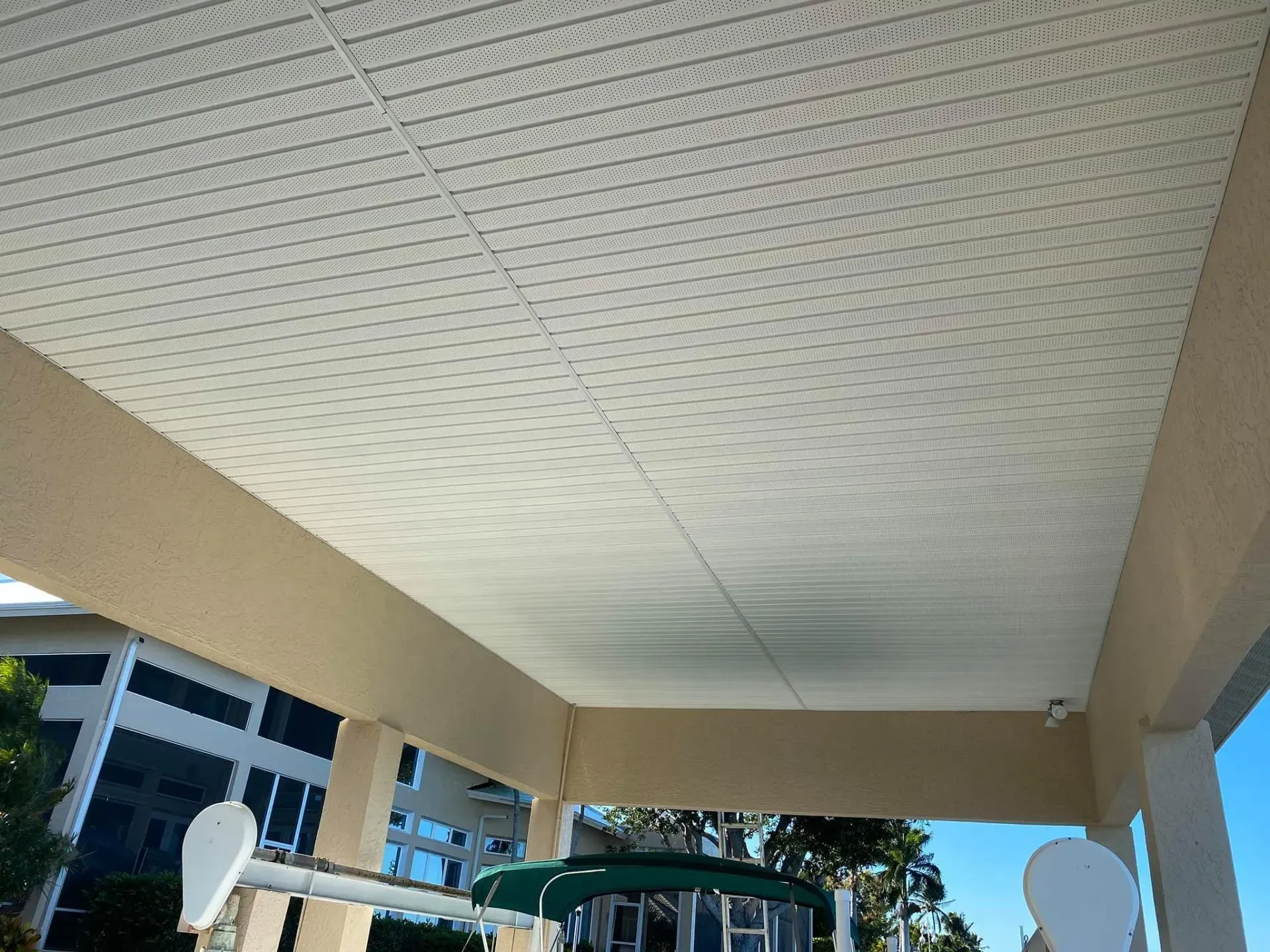 Covered walkway ceiling with beige beams and perforated white panels, leading toward a blue sky and building.