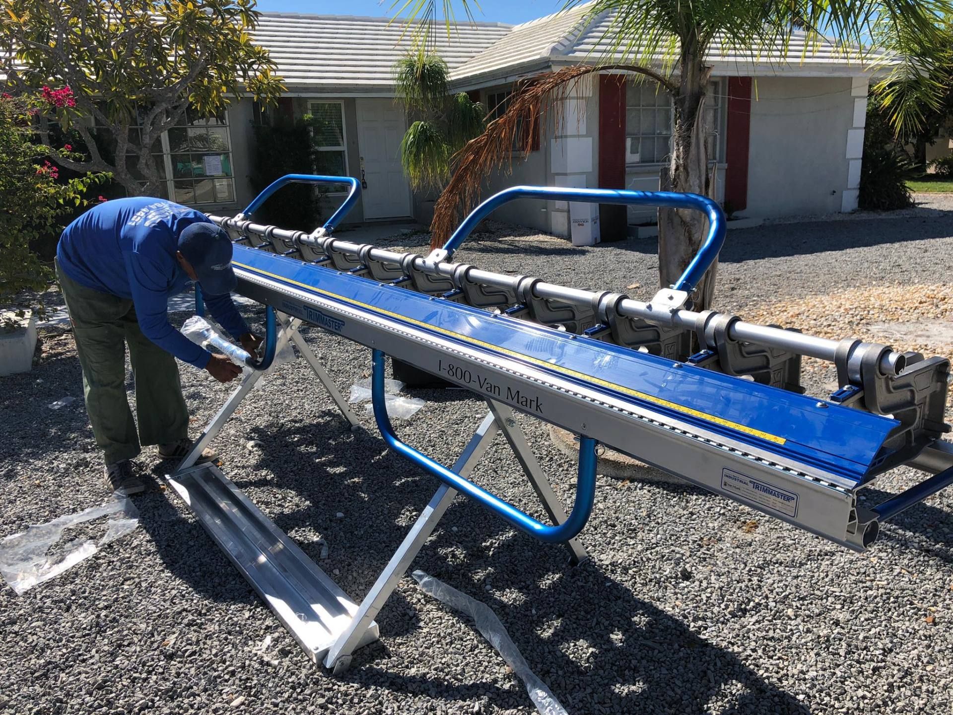 Person loading a blue metal beam onto a trailer in a driveway under a house carport.