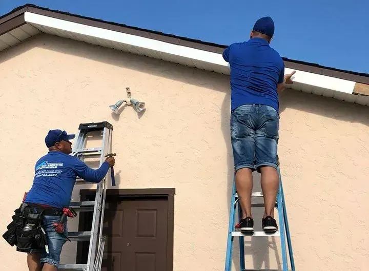 Two workers on ladders repairing a house exterior near the roofline