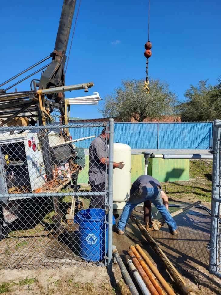 Two men are working on a water pump in a field behind a chain link fence.