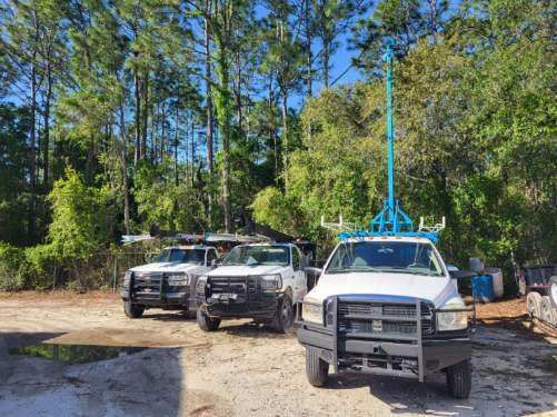 Three trucks are parked in a dirt lot in front of trees.